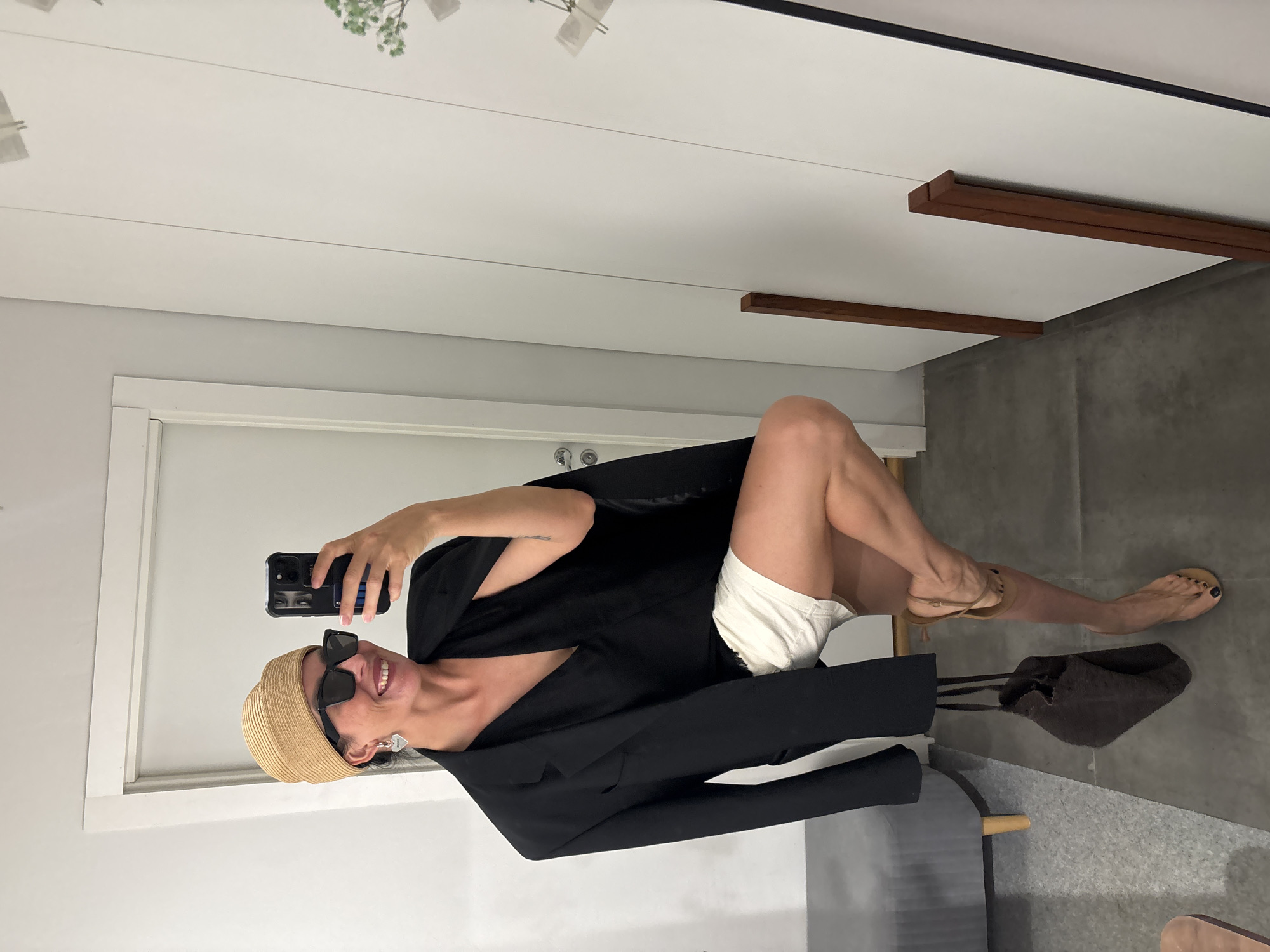 A woman stands in what appears to be a dressing room or a similar indoor space, posing for a mirror selfie. She is wearing a straw hat, black sunglasses, a black blazer draped over her shoulders, a black v-neck top, and white shorts. She is also carrying a dark brown bag and wearing tan sandals. Her left leg is bent and raised, and her right leg is extended. The background shows a white wall with vertical paneling, a white door, and a grey bench. There are no discernible elements in the image that indicate the location is the city of Chaikovsky, Russia.