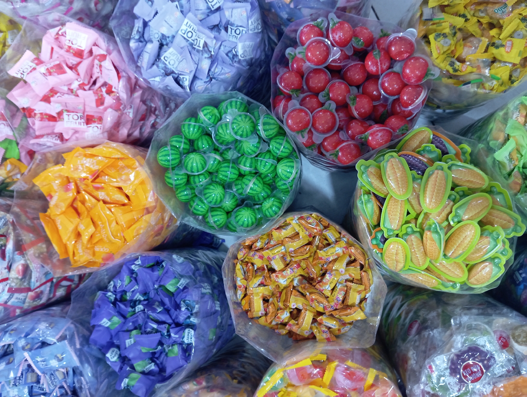A variety of individually wrapped candies are displayed in clear plastic bags, arranged in a market setting in Nha Trang, Vietnam. The bags are filled with candies of different colors and shapes, including blue, orange, pink, green, and red. Some of the candies are shaped like watermelons, corn, and tomatoes. The background shows more bags of candies and possibly shelves or other market stalls. The lighting suggests an indoor environment, possibly a candy store or a market stall. There are no people visible in the frame, and the focus is solely on the merchandise.
