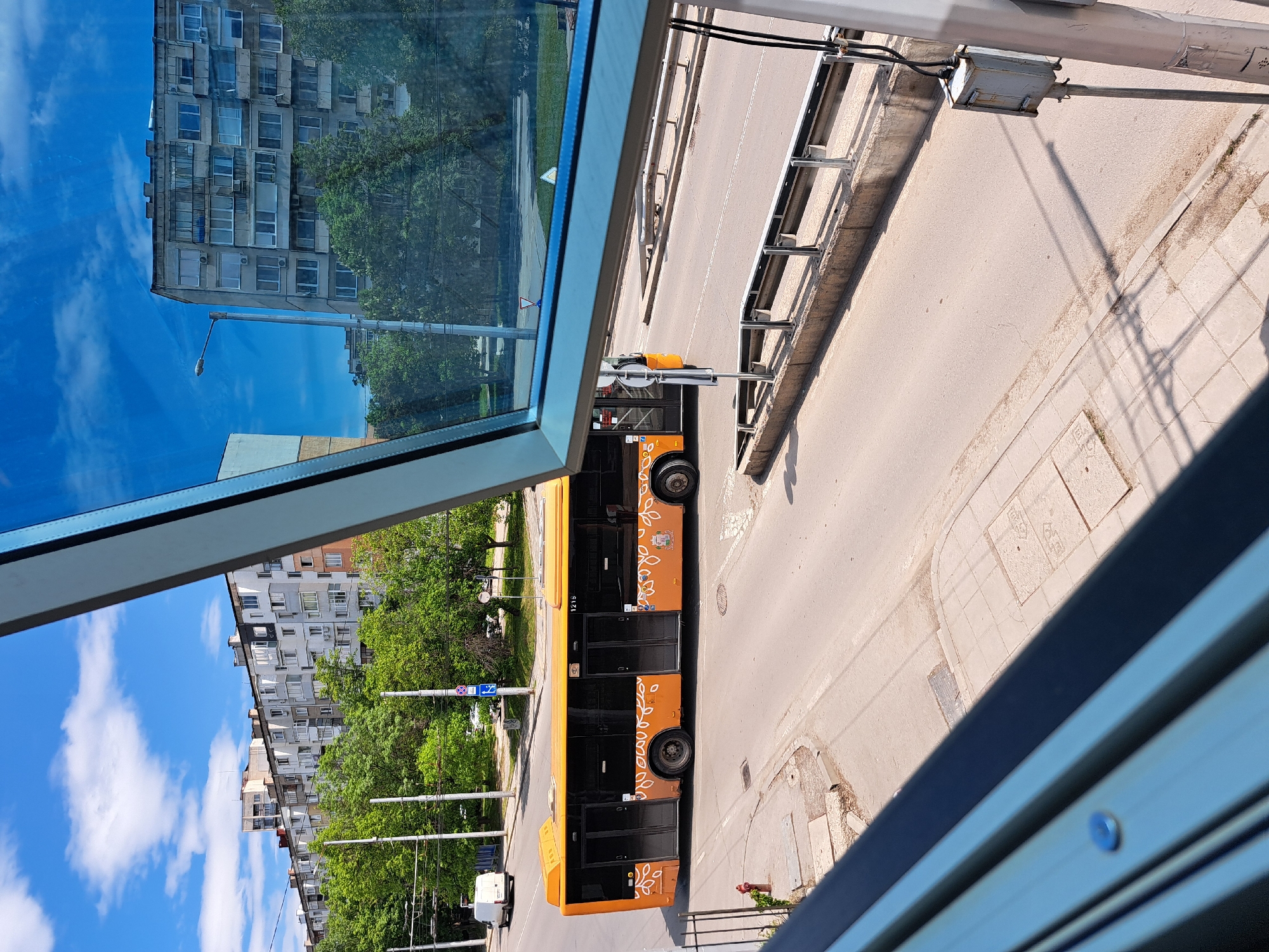 A high-angle view shows a street scene in Sofia, Bulgaria. An orange bus with floral designs and the number "1215" is parked on the road. To the left, foliage and buildings are visible. The sky is blue with scattered clouds. A reflective surface partially obstructs the view, creating a mirroring effect of a building and the sky. Overhead wires and other infrastructure elements are visible, as well as a light-colored sidewalk. The image is taken from a high vantage point, possibly from within a building or elevated structure.