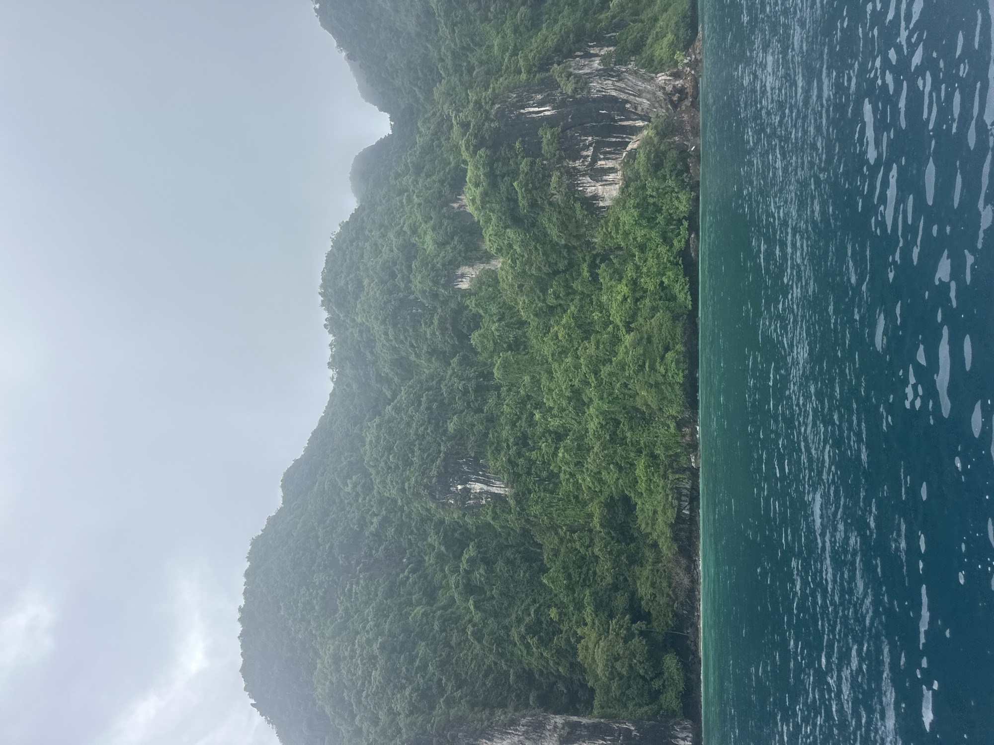 The image depicts a wide shot of a coastline with steep, jungle-covered cliffs rising from the water under an overcast sky. The water in the foreground is a deep turquoise, with small whitecaps and ripples visible. The cliffs are densely vegetated with lush green trees, and sheer rock faces with streaks of white are visible in places. The sky is a pale, diffused gray, suggesting a cloudy or foggy day. The scene appears to be a natural landscape, likely a bay or cove. There are no people or man-made structures immediately visible. The location is identified as Haad Hin Khom, Thailand.