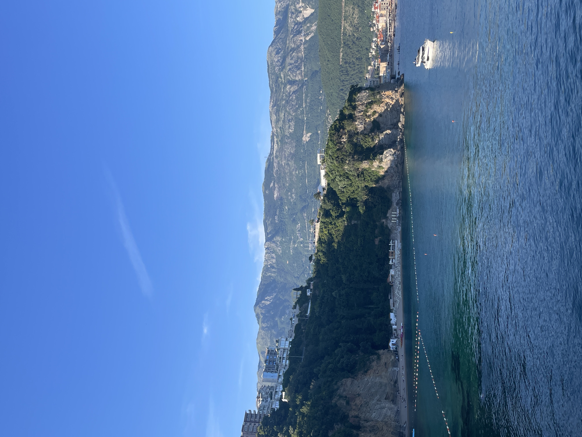 The image is a scenic shot of the coastline near Budva, Montenegro. In the foreground, a beach is lined with structures that may be hotels or apartments built into a cliffside. The beach has markers indicating a safe swimming area. Further out, the Adriatic Sea transitions from shallow turquoise near the shore to deeper blue water. A boat is visible offshore, and the landmass beyond features a town or city. A high mountain range runs along the horizon under a clear blue sky.