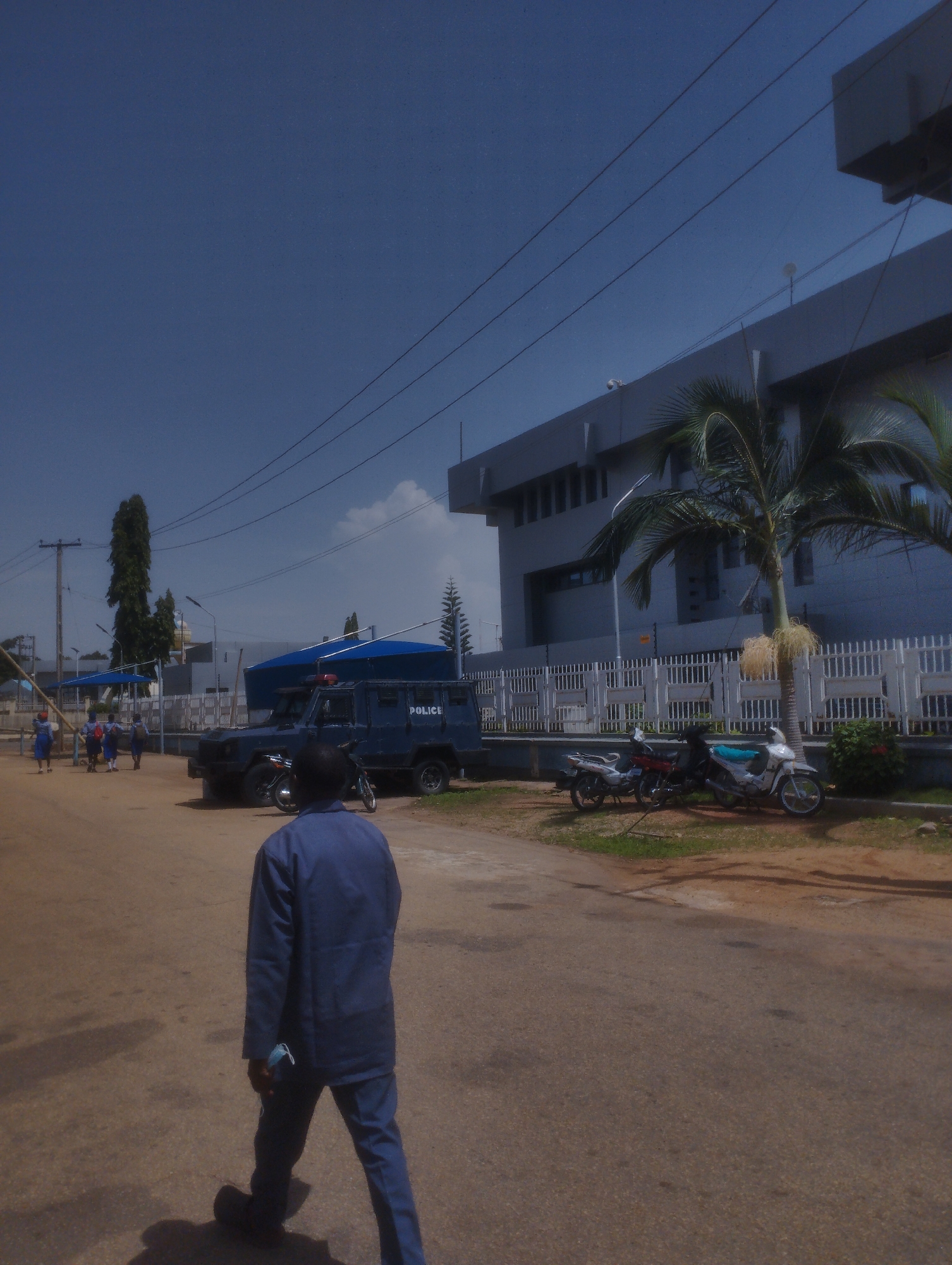 A daytime exterior shot captures a street scene in Jos, Nigeria, featuring a dark blue armored police vehicle with the word "POLICE" visible on its side. Several motorcycles are parked near a white metal fence and a large, modern building. In the foreground, a person in a blue uniform walks away from the camera. In the background, a few individuals in blue uniforms, possibly students, are seen near blue canopies and a tall tree. The sky is clear and blue.