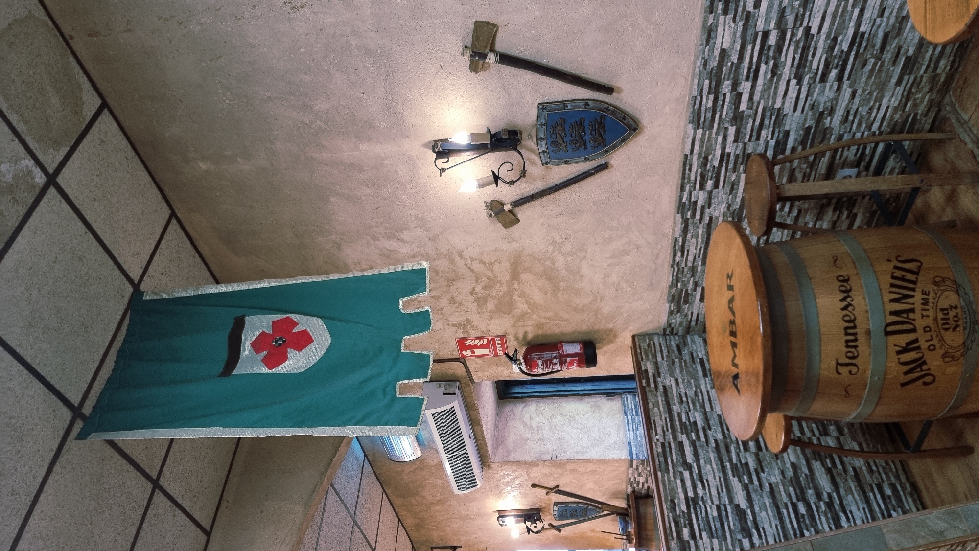 The image depicts the interior of a establishment with a rustic and thematic decor, potentially a bar or restaurant, located in the medieval town of Besalú, Spain. The walls have a textured, light brown finish. A large green banner with a silver border and a red and white shield emblem hangs from the ceiling. Below the banner, an air conditioning unit is mounted on the wall, and to its right, a red fire extinguisher is prominently displayed next to a doorway.
Further to the right on the wall, there are decorative elements including two wall sconces with candles, a shield with three lions, and two large hammer-like objects mounted on either side of the shield.
In the foreground, a table fashioned from a whiskey barrel, specifically a Jack Daniel's barrel, stands with a wooden tabletop displaying the word "AMBAR". Two wooden stools surround the barrel table. The lower part of the wall behind the table and stools is covered with a mosaic of gray and black rectangular tiles. The floor appears to be wooden planks. The overall ambiance suggests a place with a historical or medieval theme, fitting for the location in Besalú. There are no people visible in the image.