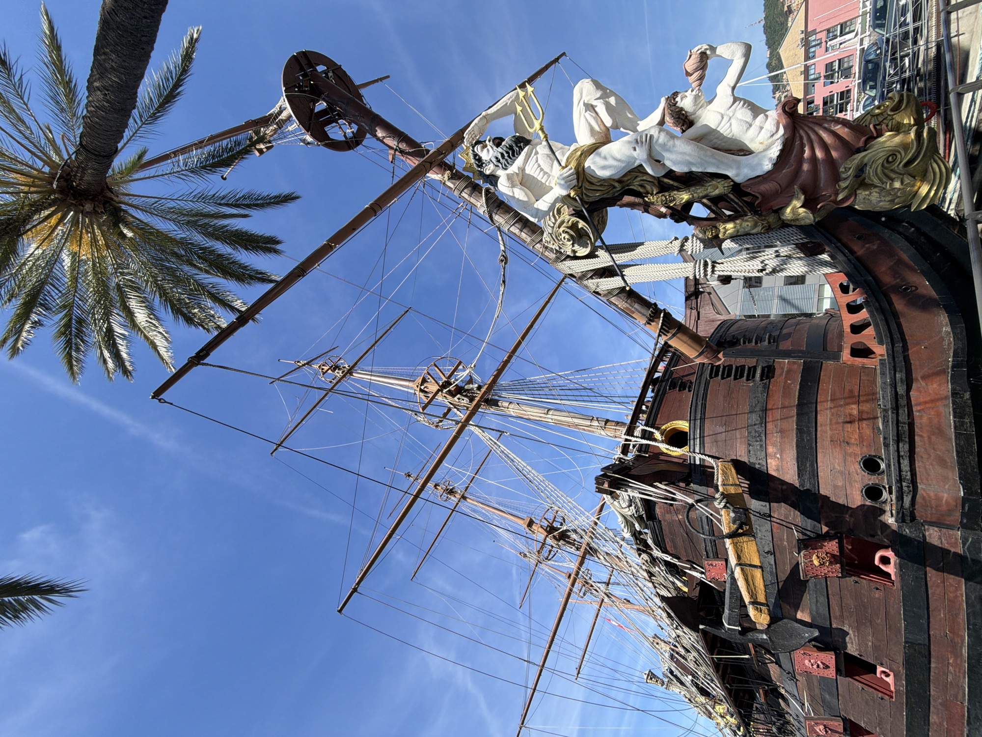 This is a low-angle shot of the Galleon Neptune, a replica of a 17th-century galleon used in Roman Polanski's 1986 film "Pirates". The ship is located in the port of Genoa, Italy. The scene features a clear blue sky with a few wispy clouds. A portion of a palm tree is visible on the left side of the image. The ship's mast and rigging extend upwards, with decorative sculptures near the top of the bow. The hull of the ship is predominantly dark brown with black stripes. Some buildings can be seen in the background.