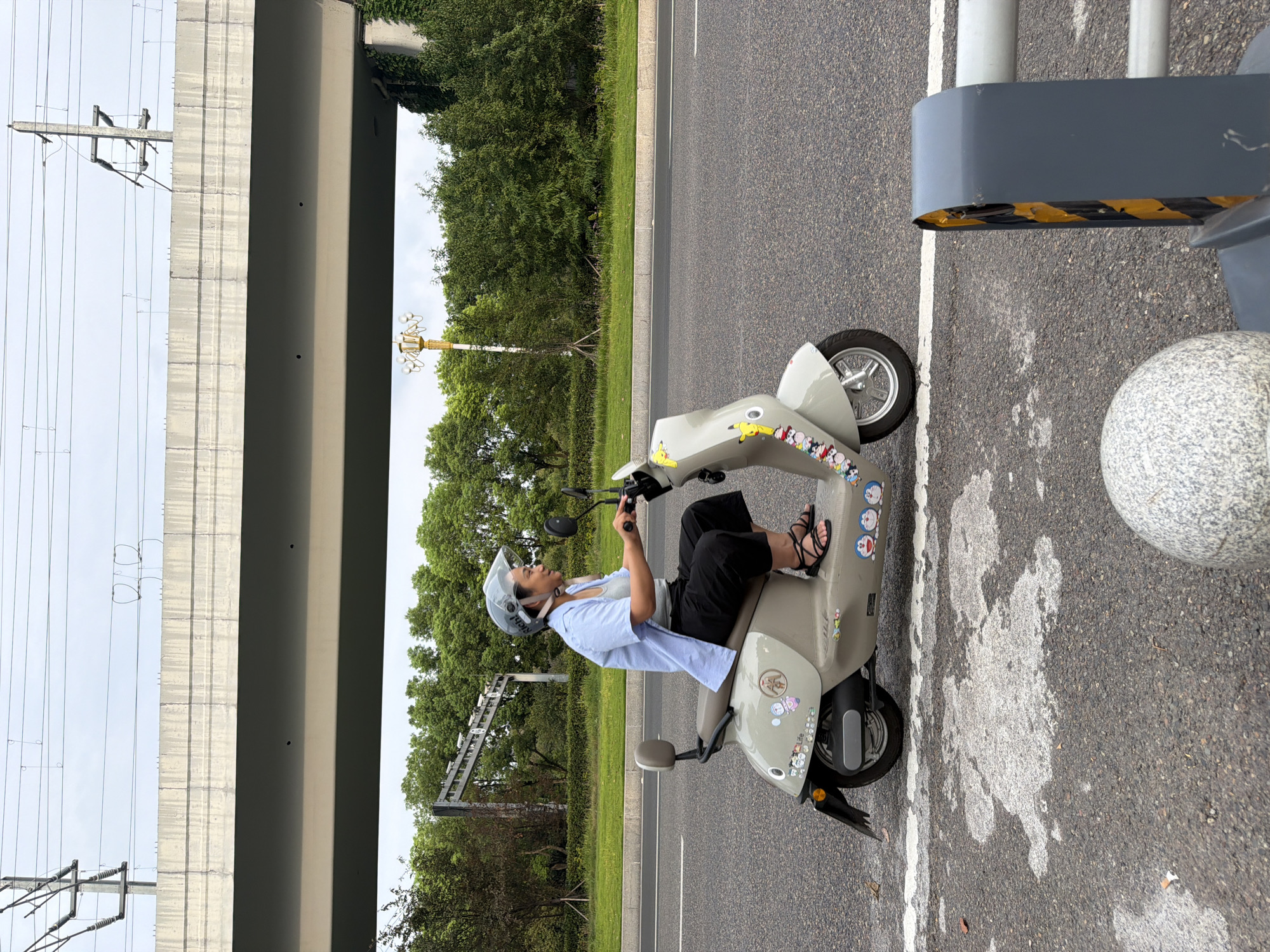 In Jiangjin, China, a woman wearing a light blue shirt, gray undershirt, black pants, and black sandals rides a light gray electric scooter. She has a clear face shield on her helmet. The scooter is adorned with various cartoon stickers, including Pikachu, Doraemon, and other characters. She is on an asphalt road with a white lane marker. In the background, there is a multi-level overpass with power lines overhead. Dense green trees line the side of the road, and a lamppost with multiple lights is visible. In the foreground, a round gray bollard and a yellow and black striped barrier are present. The sky is overcast.