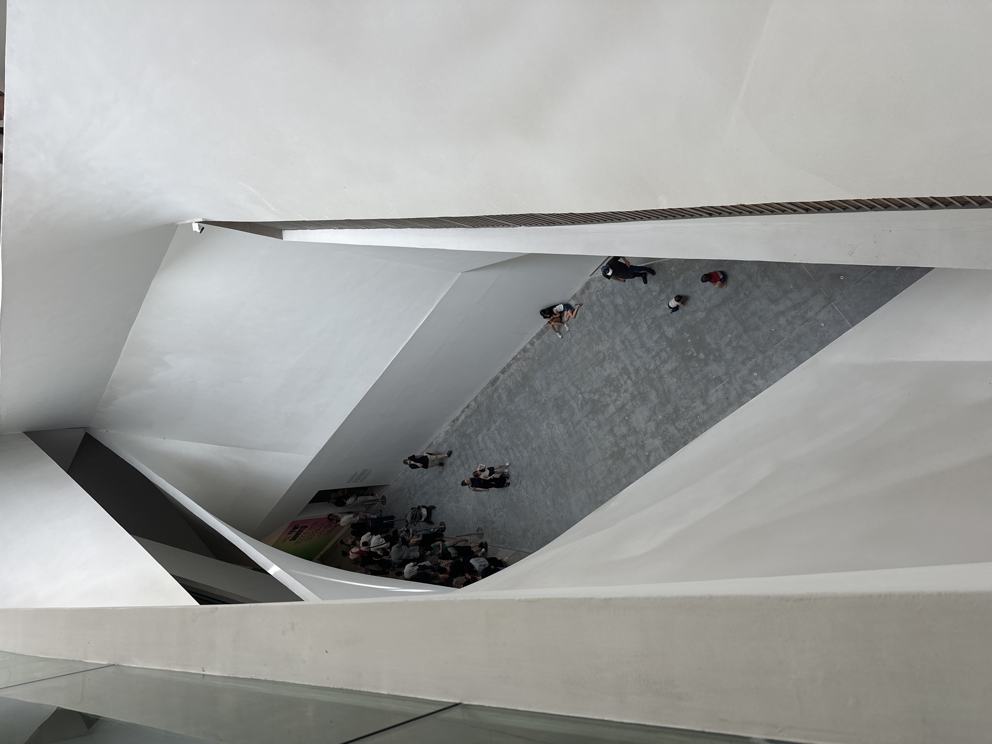 The image shows an aerial view of the interior of a building with geometric white walls. There is a group of people gathered near a poster on the lower left side of the frame. Some individuals are walking along a grey floor in the center of the image. The photograph was taken in Tel Aviv, Israel.