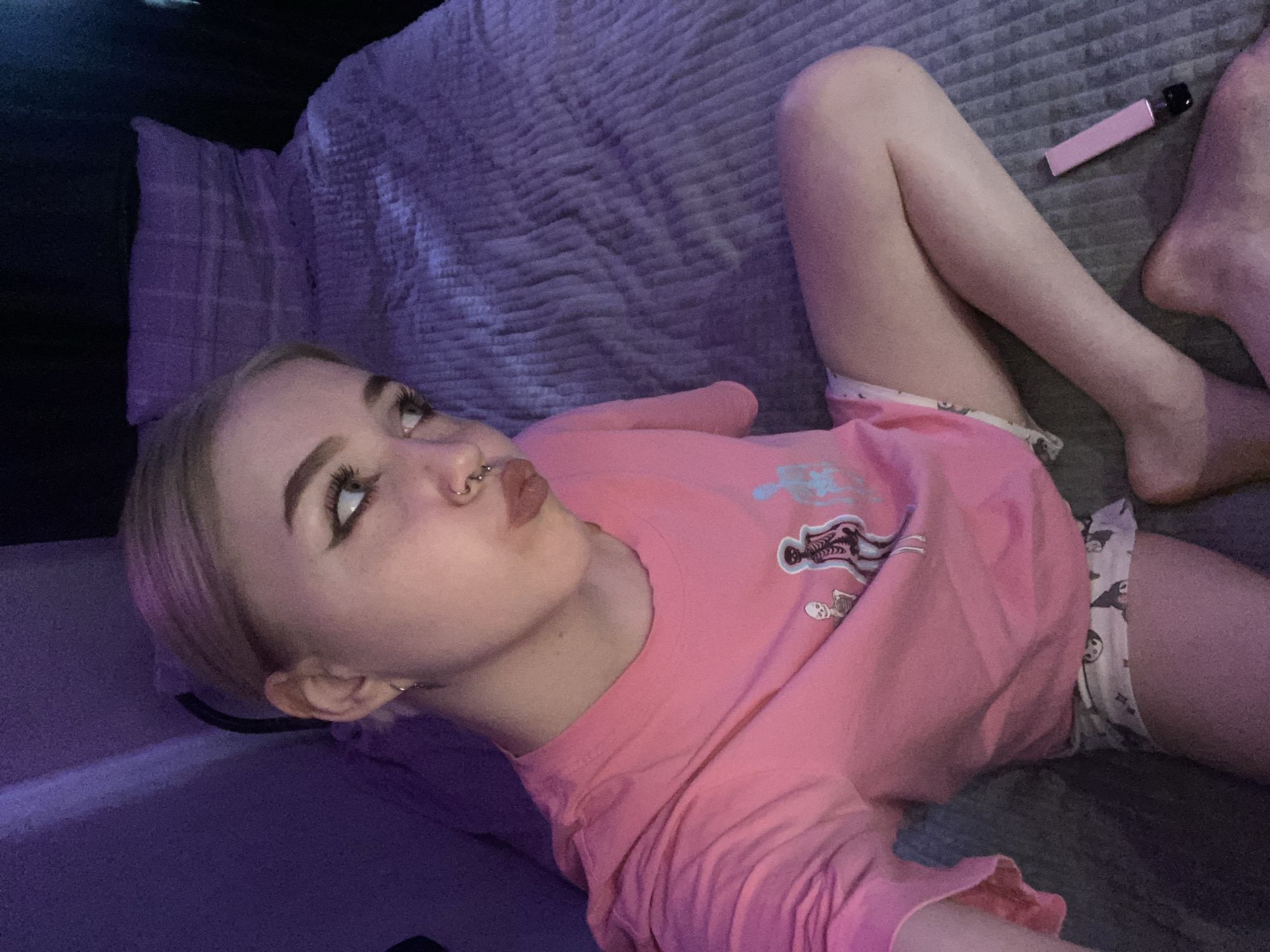 A young adult female is lying on a gray textured surface, possibly a bed, with a gray pillow behind her head. She wears a pink t-shirt with a cartoon skeleton graphic and patterned shorts. Her legs are bent, with bare feet visible near a pink object, possibly a vape pen, on the bed. The woman has fair skin, blonde hair, prominent eyeliner, and a nose ring. The environment is dimly lit with a purplish hue. There is no indication that the image was taken in Izhevsk, Russia.