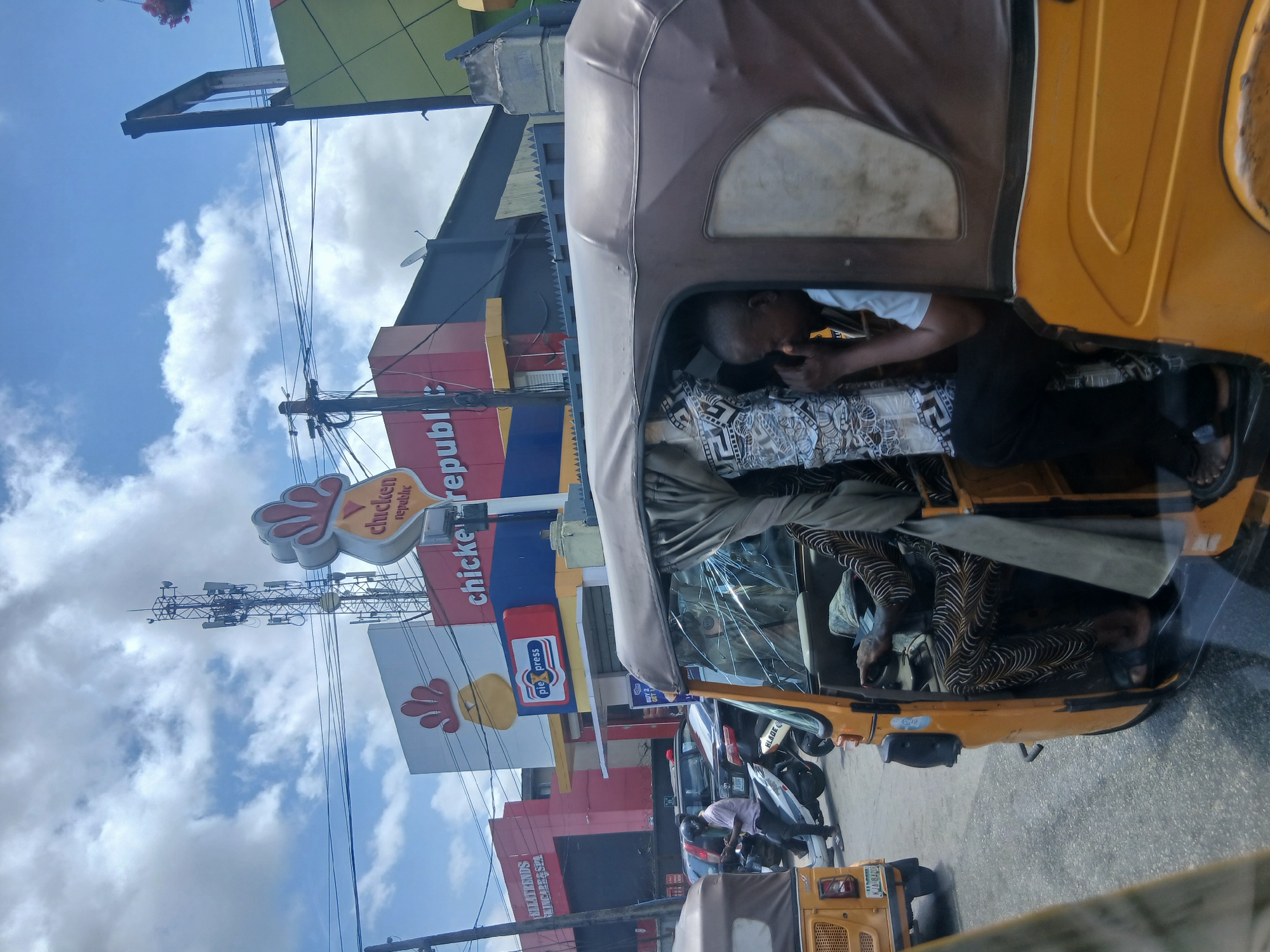 The image, captured in what appears to be Animashawun, Nigeria, shows the interior of a yellow auto rickshaw (tricycle) with three occupants. One man in a patterned shirt is partially visible in the back, while another man in a white shirt and dark pants is closer to the driver's area, possibly using a mobile phone. The exterior reveals a "Chicken Republic" restaurant with a sign including the words "pie press." Beyond the rickshaw, there's street traffic with other vehicles, including a car and more rickshaws, suggesting an active commercial area. Overhead, utility poles with multiple wires crisscross the sky, which is partly cloudy. The image seems to have been taken from another vehicle, likely while in transit.
