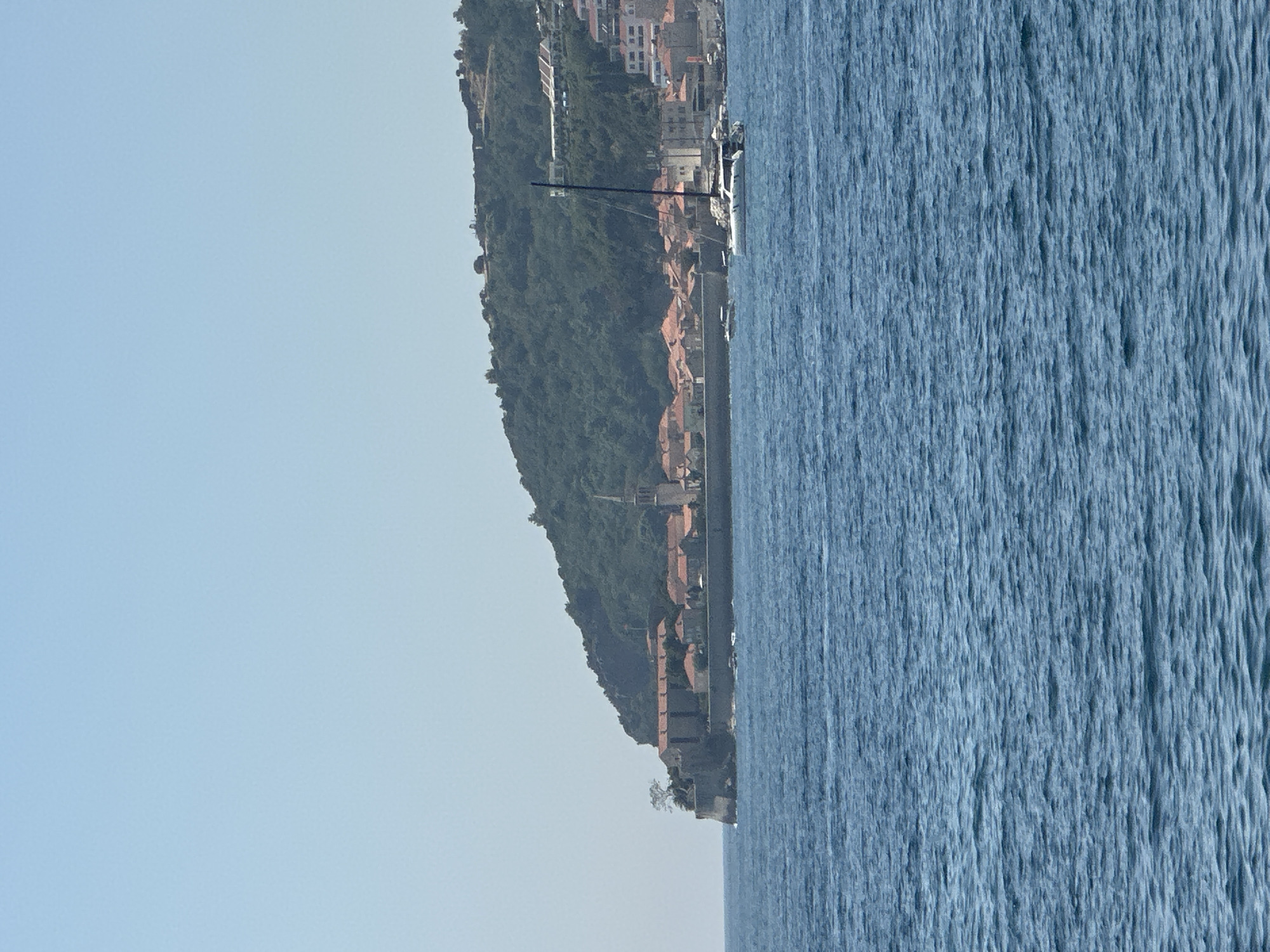 A wide shot captures a bright, clear day over the blue Adriatic Sea. In the distance, the historic Stari Grad (Old Town) of Budva, Montenegro, is perched on a peninsula jutting into the water. The fortified old town features characteristic red-tiled roofs and stone buildings, including a prominent church spire. The peninsula is backed by a lush, green hillside dotted with modern buildings and residences that climb the slope. A tall sailboat with a dark mast is anchored in the bay, alongside a smaller boat. The sea in the foreground is a deep blue with gentle ripples.