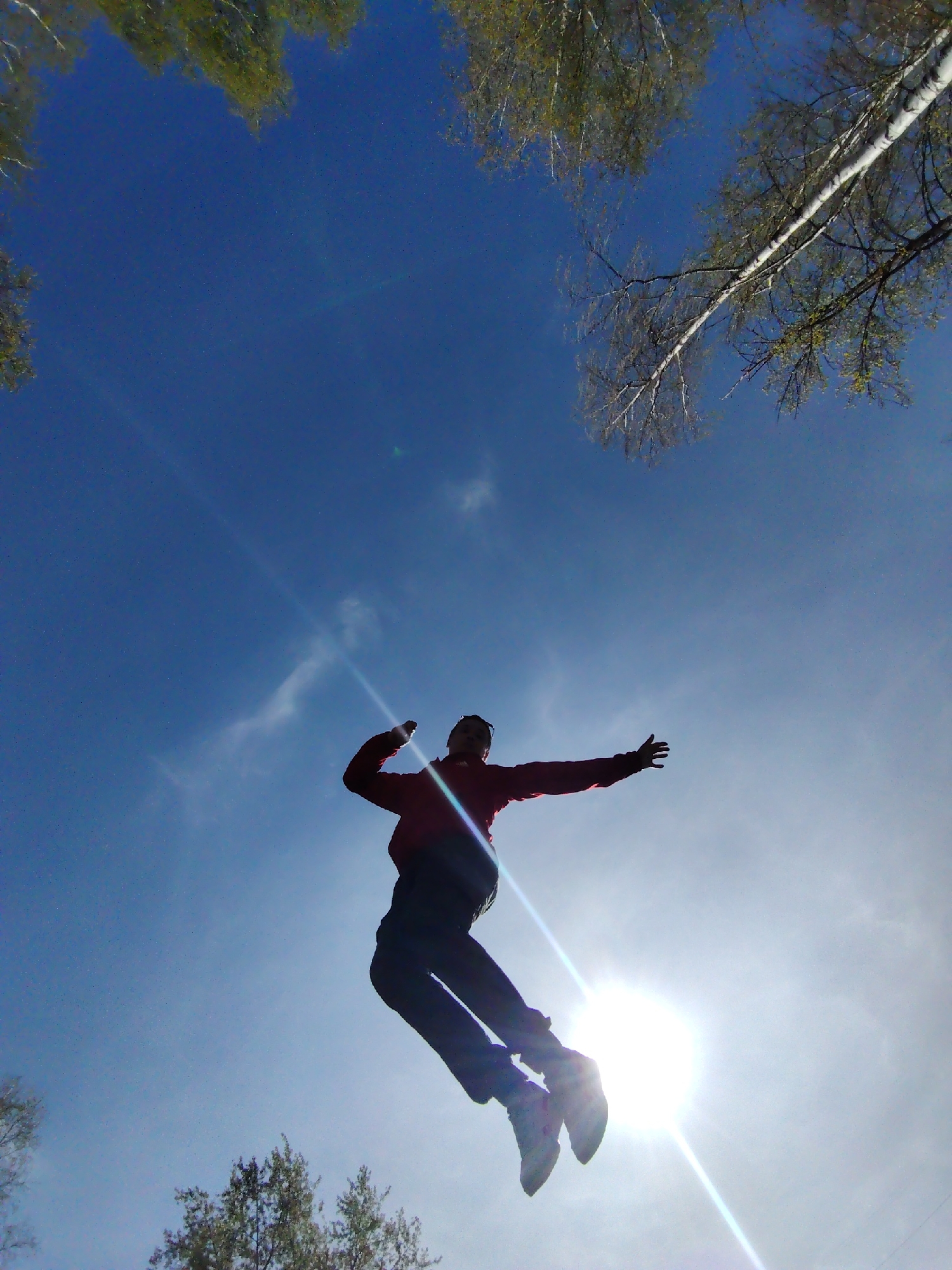 A silhouetted figure of a person is suspended in the air against a bright, sunlit sky. The person is wearing a red jacket and dark pants, with sneakers on their feet. Their arms are outstretched. The perspective is from below, looking upwards towards the person. In the upper part of the frame, branches of trees with sparse leaves are visible against the blue sky. The bright sun creates lens flare in the lower part of the frame. The picture was taken in the city of Chaikovsky, Russia.