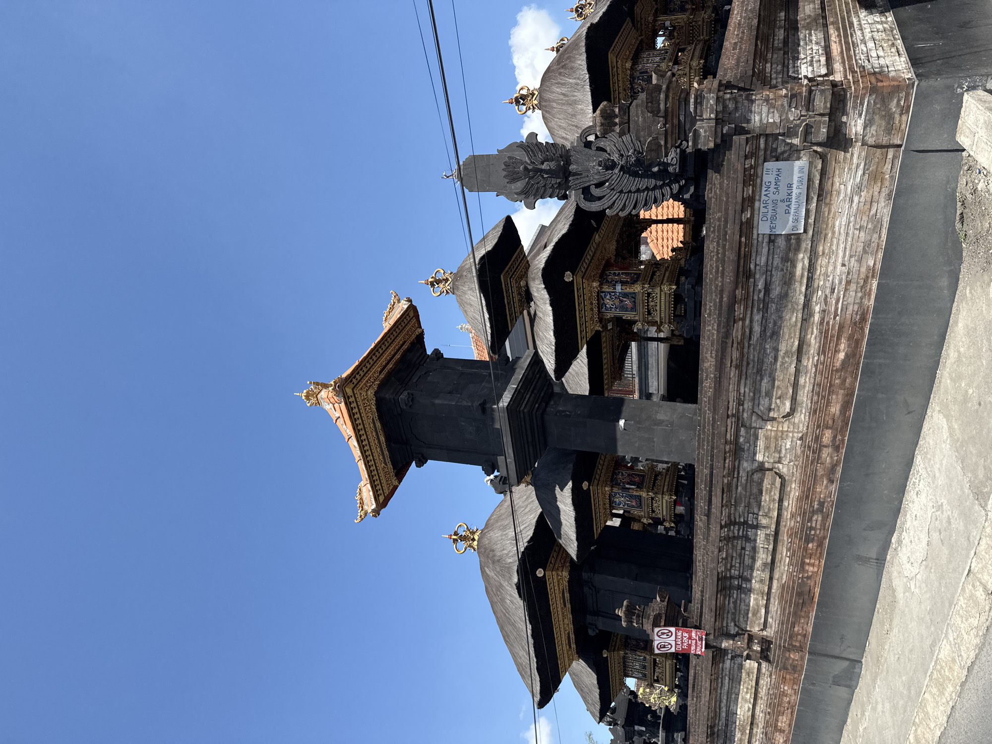 The image displays a Balinese temple structure with multiple tiered roofs topped with gold ornamentation. The temple is constructed with dark stone and intricate golden details, with thatched roofs. A tall, dark stone pillar with carved details stands prominently to the right of the central structure. A brick wall with a grey painted base runs along the foreground. Two signs are visible on the wall. One sign, red and white, has two circular symbols indicating no parking and the text "DILARANG PARKIR" and below it "PENYEBAR SAMPAH SEPALULA". A second, white sign with black text states "DILARANG !!! MEMBUANG SAMPAH & PARKIR DI SEPANJANG PURA INI". The sky is clear blue with a few white clouds. The scene is outdoors, under daylight conditions, with the camera angled upwards from street level. This is the entrance to the Jagatnatha Temple, located in Denpasar, Bali, Indonesia.