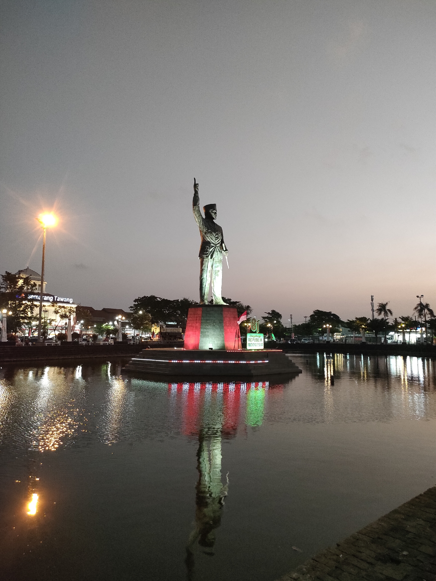 The image captures a statue of a male figure in uniform, possibly a historical leader, standing on a circular platform in the middle of a body of water. The statue is illuminated, and the platform is decorated with red and green lights. The sky is a dusky grey, suggesting it is either early morning or late evening. In the background, there are buildings, trees, streetlights, and a sign that reads "Semarang Tawang." The water reflects the lights from the statue and the surrounding environment. The overall scene appears to be a public monument in the city of Semarang, Indonesia, likely a landmark or a place of historical significance. The statue is the central subject, with its arm raised and pointing upwards. There are no visible people in the image.
