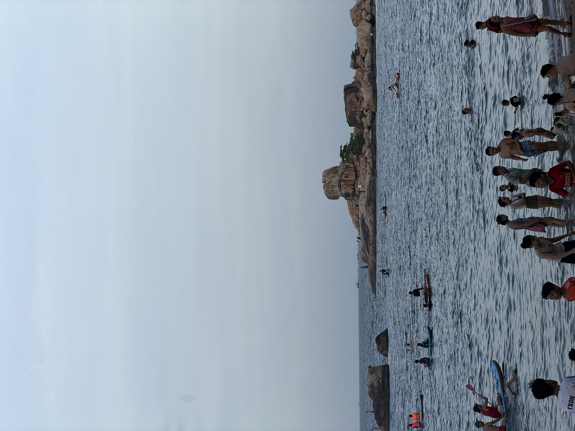 A group of people are enjoying a day at the beach in Bac Nha Trang Ward, Vietnam. The sky is overcast, and the sea is calm with small waves. In the foreground, several people are standing in the shallow water, while others are swimming or paddleboarding further out. In the middle ground, a cluster of large rocks and smaller islands form a rocky outcrop. Several individuals are paddleboarding or kayaking around these rocks. In the background, a few boats are visible on the horizon. The beach itself is sandy, and there are scattered rocks along the shoreline. The overall scene depicts a leisurely day at a popular coastal destination.