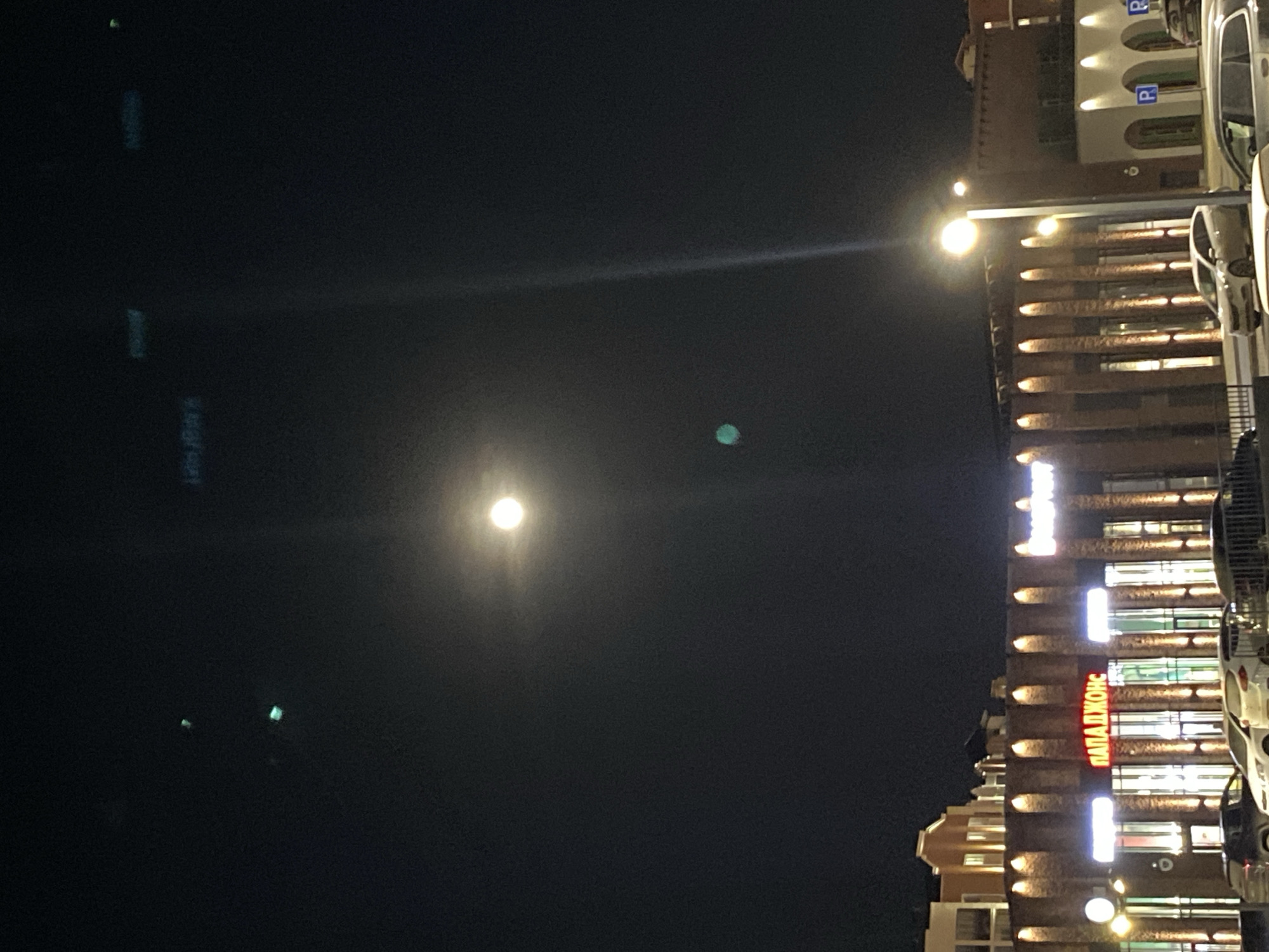 The image shows a night scene in Izhevsk, Russia. The moon is visible in the dark sky with a bright halo. A building is illuminated with multiple lights, and a sign in Russian script that translates to "Papa John's" is visible. Several parked vehicles are present, and a street lamp is lit. The location is likely near the Papa John's restaurant in Izhevsk. No people are visible in the image.
