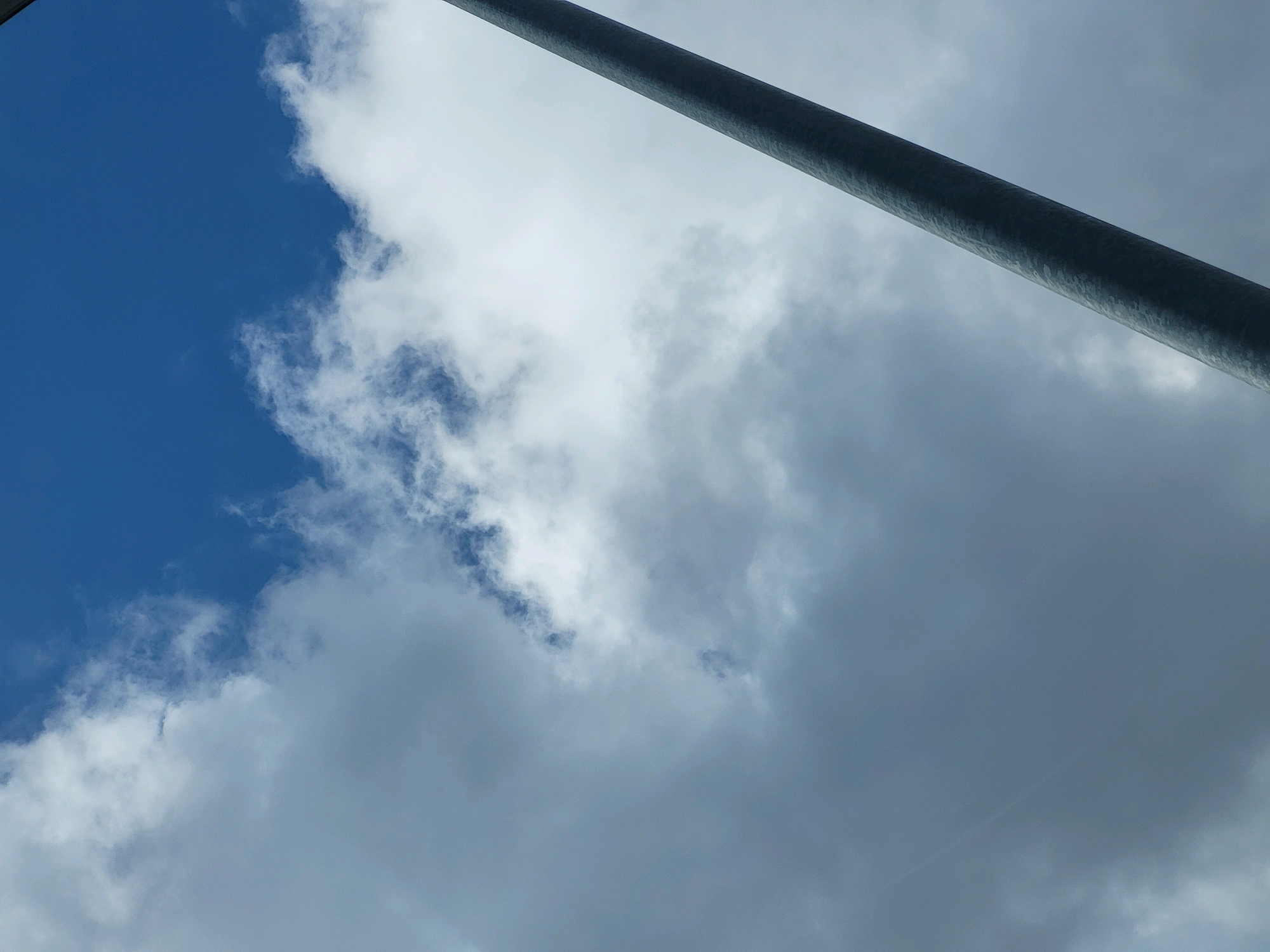 A tall, slender, dark grey metallic pole runs diagonally across the upper right portion of the image, from the top edge downwards and towards the left. The rest of the frame is filled with a cloudy sky. On the left side, patches of bright blue sky are visible, interspersed with wispy white clouds. As the frame moves towards the right, the clouds become denser and darker, transitioning to a uniform grey sky. The image appears to be taken from a low angle, looking upwards. The location is not specified in the image or provided context.