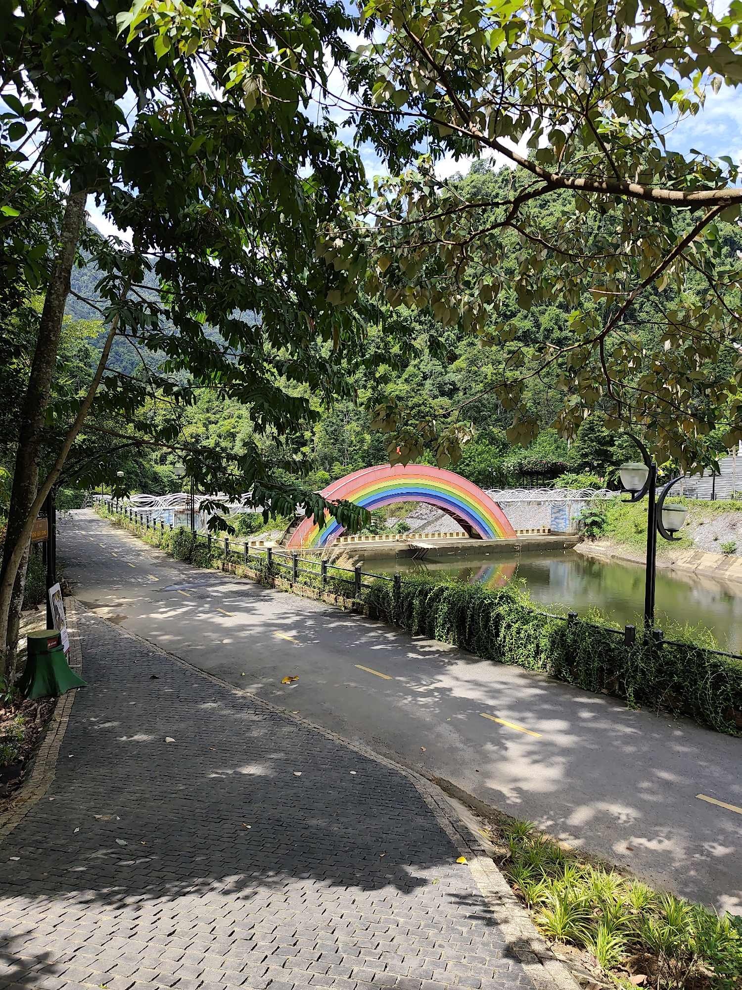 An outdoor scene shows a paved walkway and road next to a body of water, with a vibrant rainbow arch bridge spanning across it. The walkway on the left is made of dark grey cobblestones, while the road is asphalt with a yellow line down the center. Lush green trees with dappled sunlight cast shadows on the ground. The water reflects the sky and surrounding greenery. The rainbow bridge is the prominent focal point, featuring distinct bands of red, orange, yellow, green, blue, and purple. Along the edge of the water is a fence with green vines growing on it. Two lampposts with white globes stand near the road. In the background, a dense forest covers a hillside under a partly cloudy sky. The image captures a serene and colorful park-like environment.