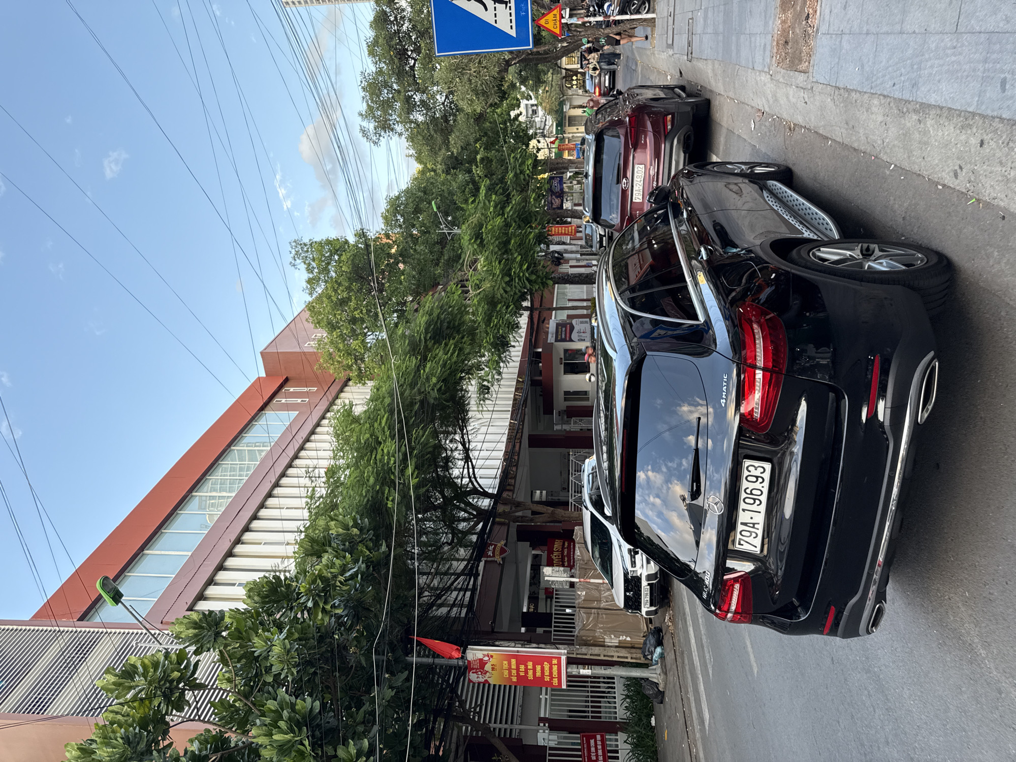 The image depicts a street scene in Nha Trang, Vietnam. In the foreground, a black Mercedes-Benz GLC 300 with license plate 79A-196.93 is parked facing away from the camera. To its right, a burgundy Hyundai Santa Fe with license plate 79A-248.02 is also parked. Further down the street, other vehicles and pedestrians are visible. The street is lined with trees and a modern building with red and white accents. Overhead, numerous electrical wires crisscross the sky. A pedestrian crossing sign and a yellow warning sign are present on the right side of the image. The overall atmosphere suggests a typical urban environment on a clear day.