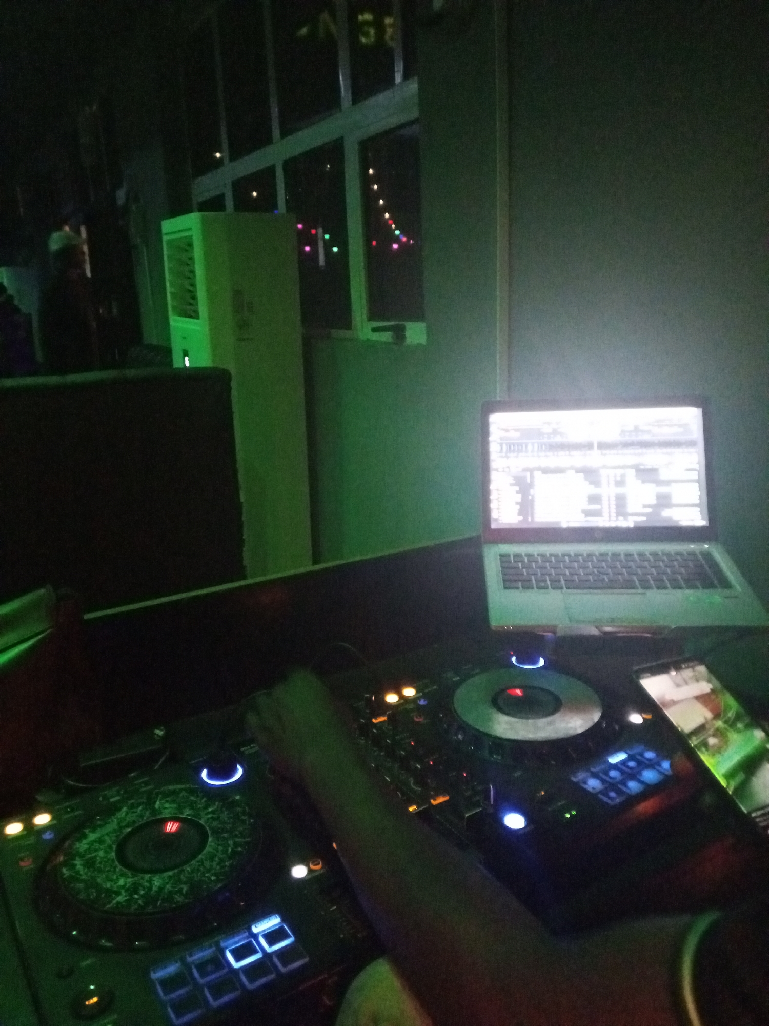 The scene shows a DJ setup in what appears to be a dark nightclub or bar. The lighting is dim and dominated by green hues. The DJ setup includes two turntables, a mixer, a laptop with music software displayed on the screen, and a smartphone. A person's hand is visible on one of the turntables, suggesting someone is actively DJing. Background elements include a window and an air conditioning unit. The image was taken in Lokoja, Nigeria.
