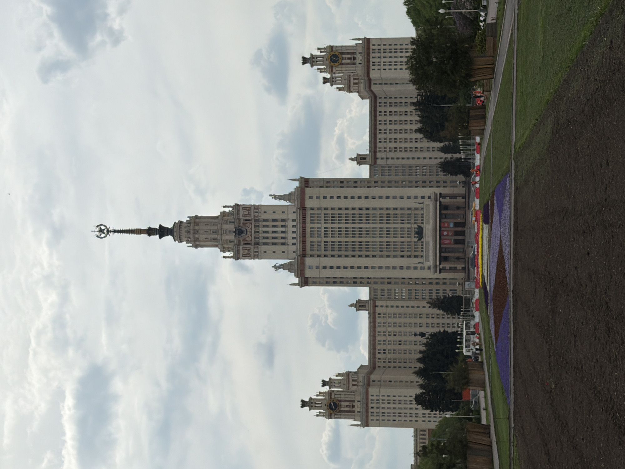 The image shows the Moscow State University building in Moscow, Russia. The main building is a tall skyscraper with a spire and a star on top. It has a symmetrical design with many windows. The building is surrounded by green grass and trees. In the foreground, there are flowerbeds and paths. The sky is cloudy.