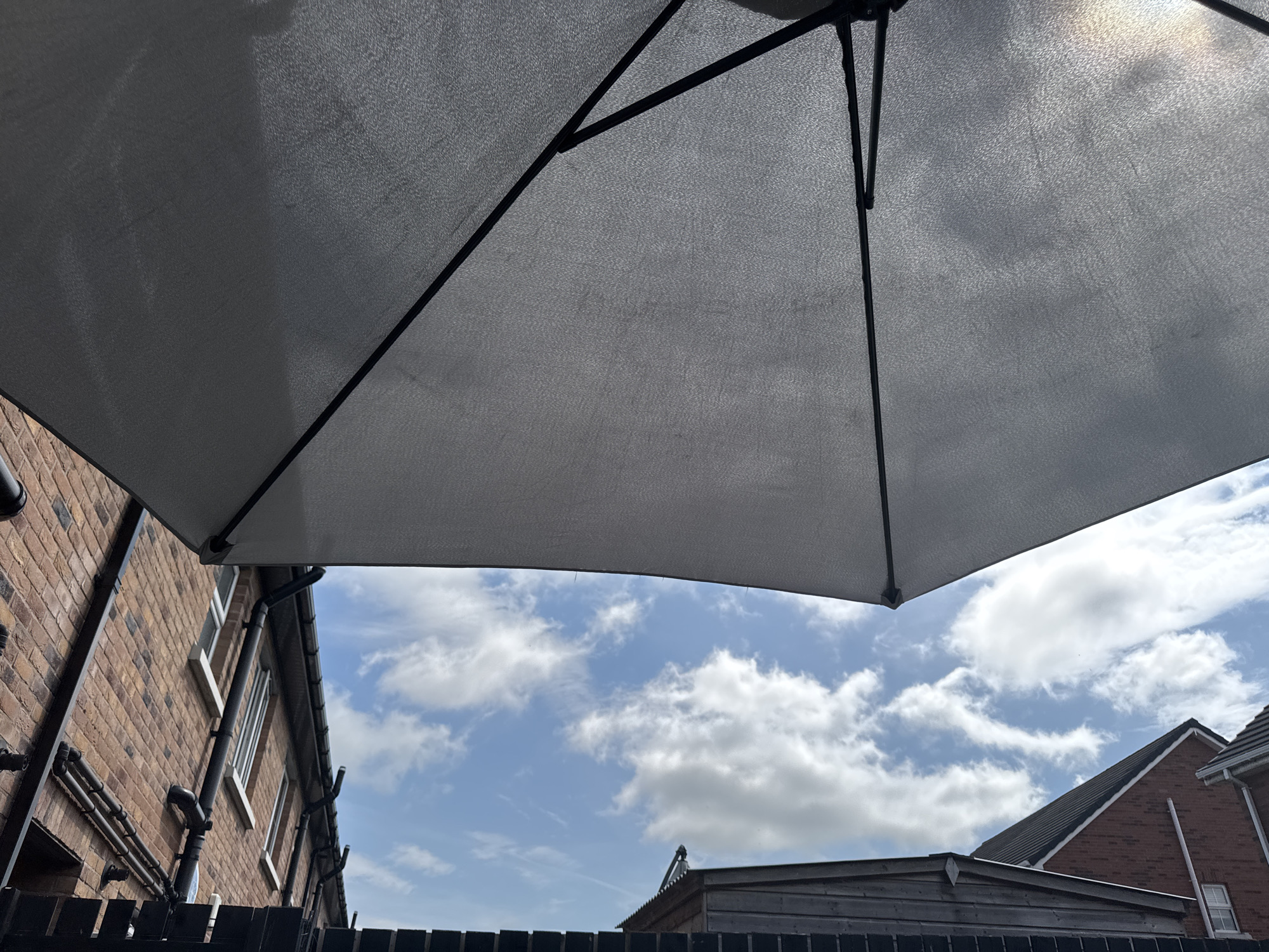 The image captures an outdoor scene from a low-angle perspective, looking upwards. The foreground is dominated by the underside of a large, light-grey umbrella, supported by black metal poles. The fabric of the umbrella shows a textured weave and is illuminated by sunlight, creating some dappled light effects.
Beyond the umbrella, a bright blue sky is visible, dotted with fluffy white clouds. The setting appears to be a residential area, with architectural elements of houses visible. To the left, a brick wall of a building with multiple windows and downpipes runs diagonally across the frame. In the mid-ground, a wooden structure, possibly a shed or outbuilding, with a dark roof is situated. To the right, the roofline and upper walls of another brick house are visible. A black fence runs along the bottom of the image, partially obscured by the umbrella's pole.
The lighting suggests it is daytime, likely midday or early afternoon, given the bright sky and the quality of light. The weather appears to be fair, with a mix of sun and clouds. There are no people or discernible activities taking place. The overall mood is one of a peaceful, sunny day outdoors. There is no visible text in the image. The provided location context of Craigavon, United Kingdom, helps to place the scene geographically.