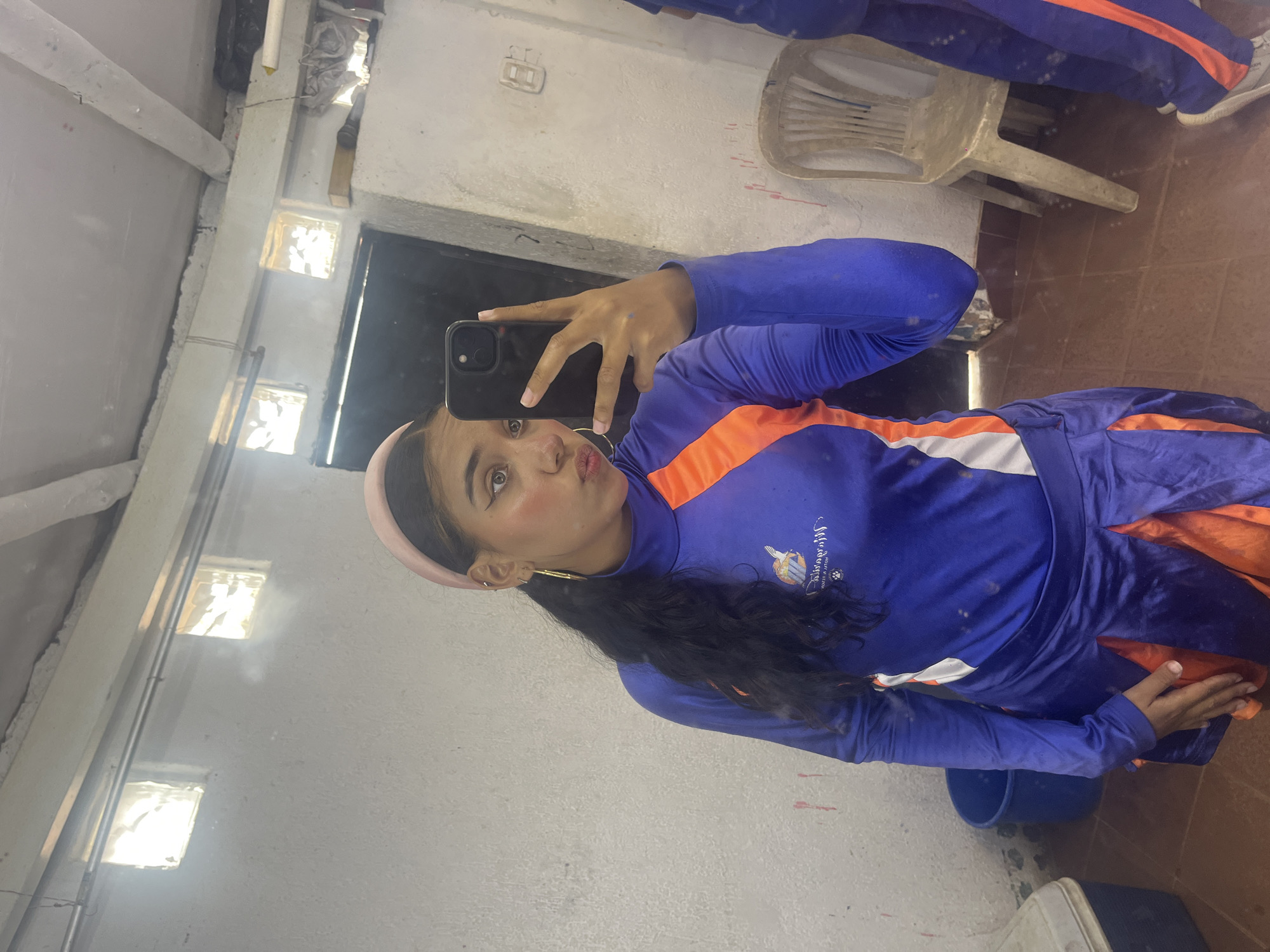 A young woman, likely of South Asian or mixed ethnicity, captures a mirror selfie in an interior space located in Porlamar, Venezuela. She has long, dark, wavy hair styled in a low ponytail and wears a pink headband. Her makeup includes winged eyeliner and blush, and her left ear displays multiple piercings, including a prominent gold hoop earring. She holds a black smartphone with her right hand, partially obscuring the lower right side of her face, and her lips are slightly pursed.
She is dressed in a two-piece athletic uniform: a royal blue long-sleeved top with orange and white vertical stripes on the chest and a logo depicting a bird, text (possibly "Melgarejo"), and a paw print. The top is paired with a matching royal blue skirt or skort featuring orange pleated panels. Specks of dirt or paint are visible on her clothing and skin.
The reflected environment reveals a utilitarian room with rough, light-colored walls, displaying reddish-brown splatters and streaks, particularly on the lower right. The ceiling features exposed concrete beams. Two small, square glass block windows positioned high on the left wall emit bright light. A blue plastic bucket and a white and blue cooler are present on the left side of the floor. To the right, a beige plastic chair with a slatted back is visible, along with a white wall outlet and miscellaneous items on a ledge above. A second individual's legs, dressed in similar blue and orange athletic attire, are partially visible on the far right. The floor is covered with terracotta-colored tiles, showing signs of dust or debris.