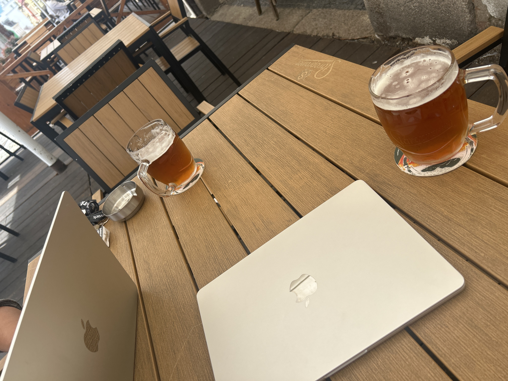 This high-angle shot captures an outdoor cafe setting, likely in Liberec, Czechia, during bright daylight. The primary subjects are a light brown slatted wooden table, two Apple MacBooks, two glass beer mugs filled with a golden-brown beverage topped with foam, a metal ashtray, and a set of keys.
The table features a faint, embossed "Staropramen" logo, a Czech beer brand, which is also visibly printed on both beer mugs. One mug is nearly full and rests on a colorful coaster with indistinct graphics, while the other is about half-full.
Two silver-colored MacBooks are open on the table, indicating ongoing use. One laptop's screen is slightly visible, while the other's lid faces up, prominently displaying the Apple logo. Next to the laptop on the left, a set of keys with a lanyard partially showing the word "work" is placed next to a small, circular metal ashtray.
In the background, out-of-focus wooden tables with dark metal frames and chairs suggest a larger outdoor dining or seating area. The ground beyond the table appears to be a paved surface. The scene conveys a relaxed atmosphere, potentially for remote work or leisure, enjoying a beer outdoors.