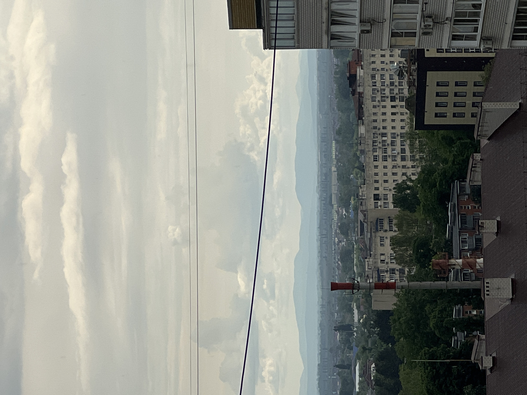 The image shows a cityscape, likely taken from a high vantage point such as a rooftop or upper floor of a building. The sky is overcast with visible clouds. In the distance, there are hazy mountain ranges. A city sprawls below, with various buildings ranging from apartment blocks to industrial structures. A tall red and white chimney is visible amidst the buildings. The scene appears to be in Krasnodar, Russia. Power lines are strung across the foreground. Buildings with varying architecture are mixed with trees.