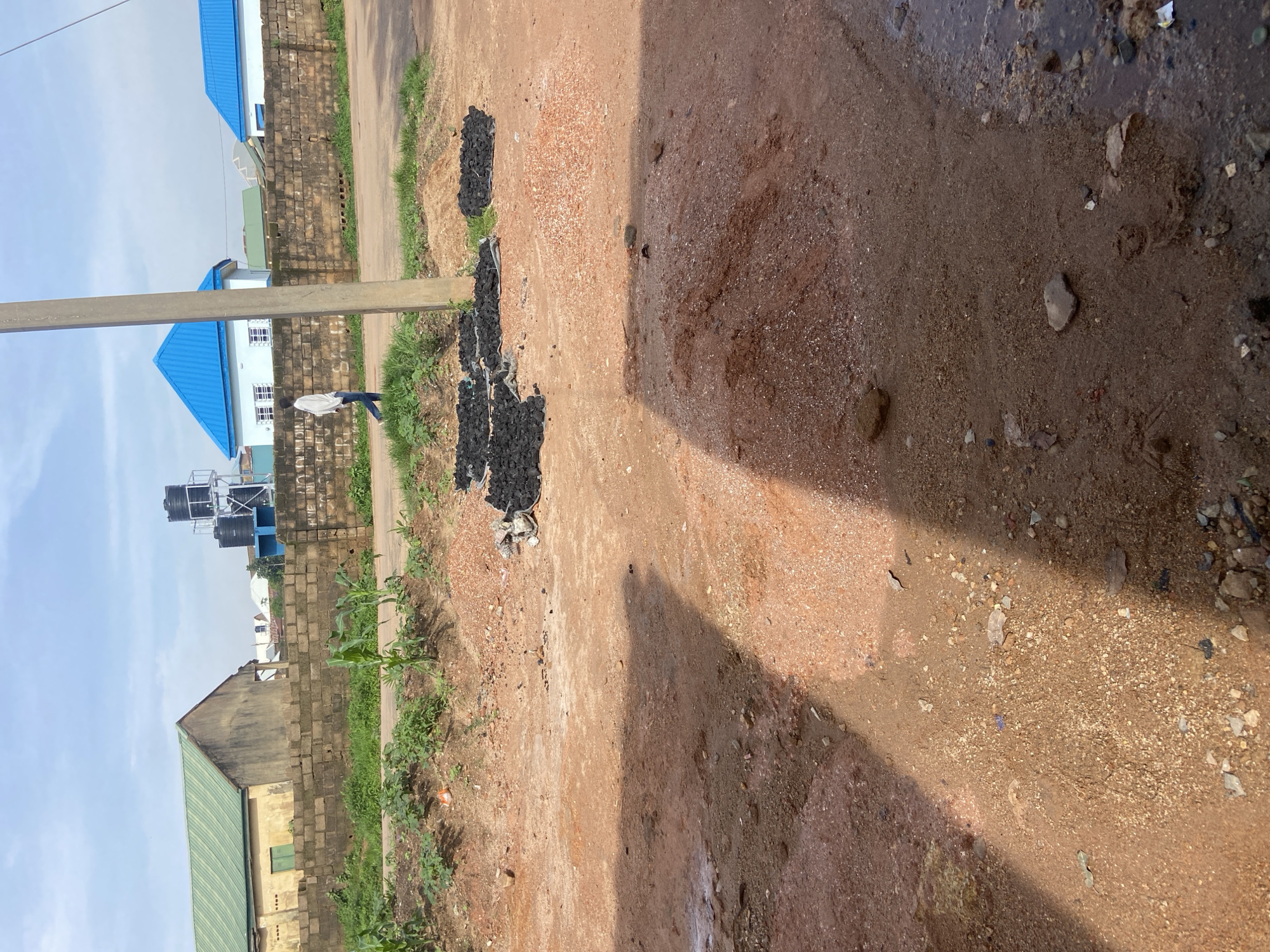 The image depicts a street scene in Jos, Nigeria, characterized by a dirt road in the foreground with piles of reddish-brown soil and gravel. Several heaps of dark, charcoal-like material are present on the side of the road, some on what appear to be white tarpaulins. A person, dressed in a white long-sleeved shirt and blue jeans, is walking away from the camera on the road, near a tall concrete pole. In the background, a low, brick wall separates the road from a strip of green vegetation and then buildings. One building has a green corrugated roof, while others feature bright blue corrugated roofs. Water tanks are visible on a raised platform behind the brick wall, near the building with the green roof. The sky is partly cloudy. The overall impression is of a somewhat unpaved urban or semi-urban environment.