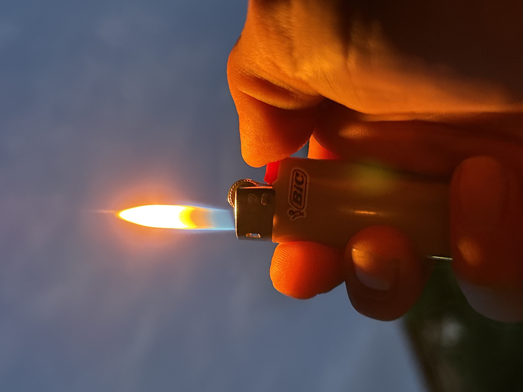 A single human hand holds an ignited orange or yellow BIC brand disposable lighter against a muted blue-grey sky with faint cloud formations. The lighter emits a bright, vertical flame, presenting a white-yellow core transitioning to orange, with a faint blue base. The flame illuminates the lighter and the fingers of the hand with a strong orange and red hue, highlighting skin textures and creases. Portions of a dark, out-of-focus background, possibly a building or foliage, are visible in the lower-left. The scene is set outdoors in the city of Unknown, Unknown, under low ambient light conditions.