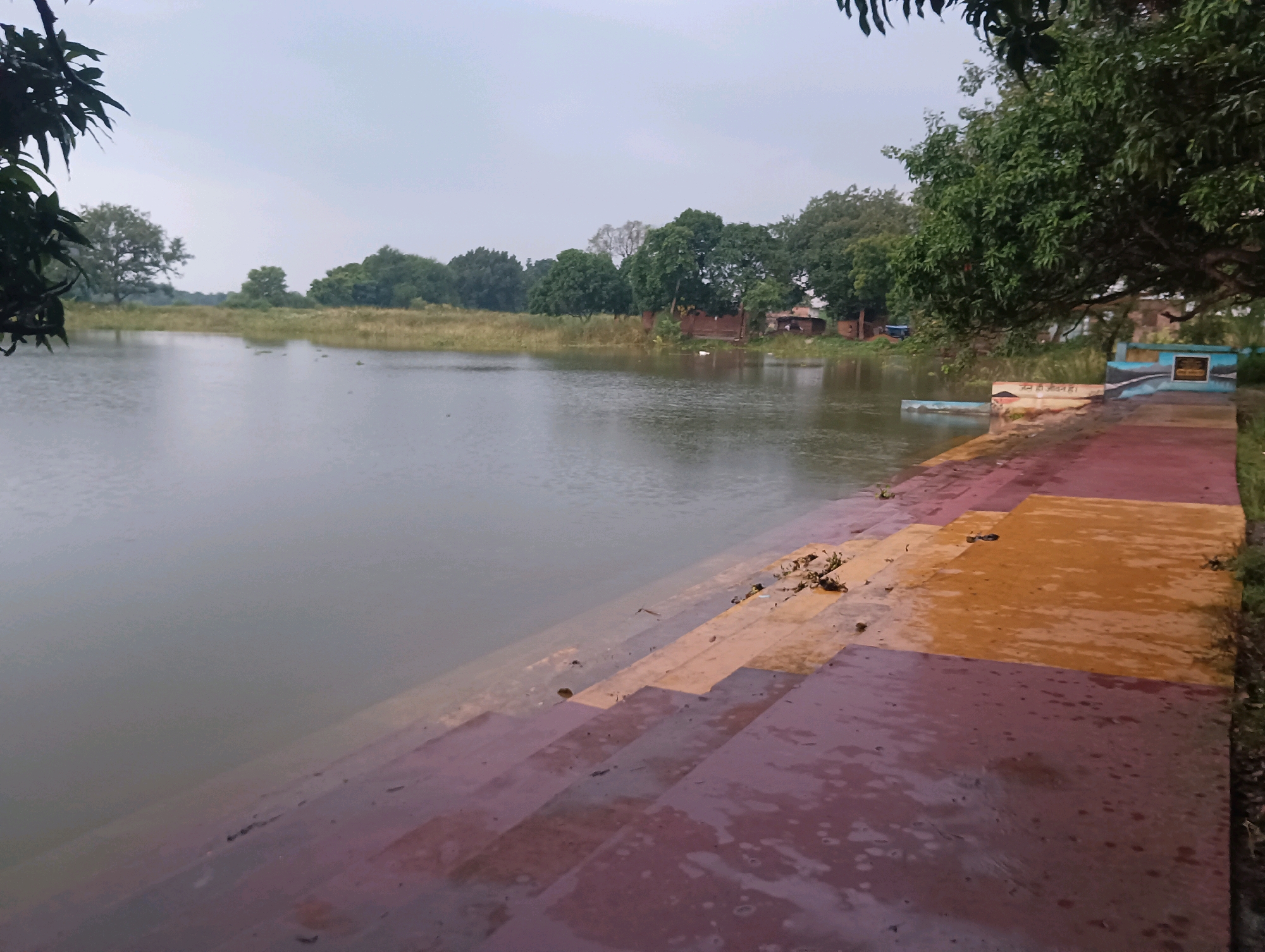 The image depicts a body of water, likely a pond or lake, on an overcast day. In the foreground, a set of wide steps, constructed from reddish-brown and orange materials, descends into the water. Some of these steps are partially submerged. To the right of the steps, there is a paved area with colorful murals featuring blue and white wave-like patterns, as well as an inscription. Vegetation, including trees and grass, is visible along the bank and in the background. The background reveals a line of trees and some small buildings, suggesting a semi-rural or suburban setting. The overall atmosphere is calm and somewhat muted due to the cloudy sky. This scene is located in Tilkar, India.