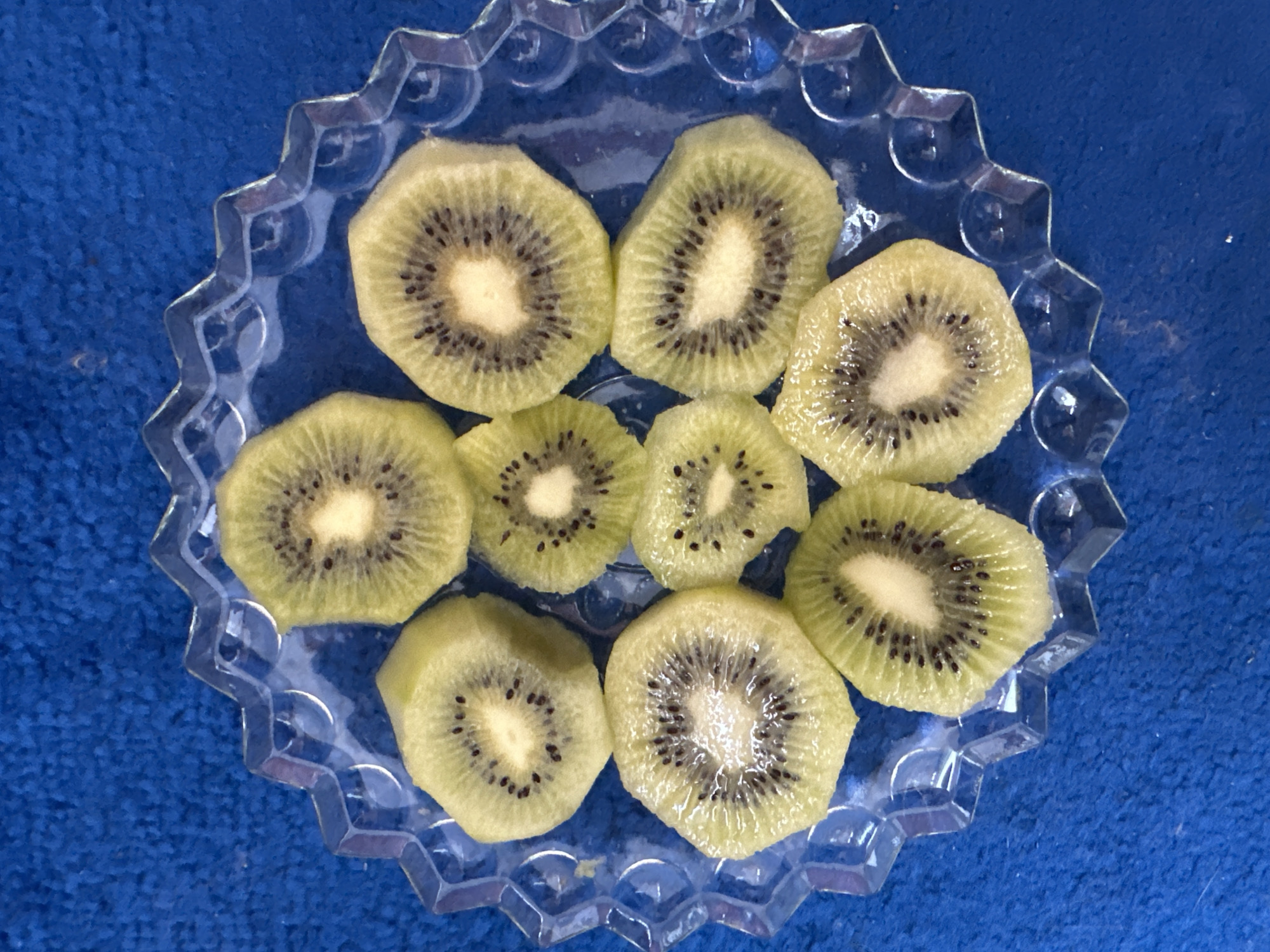 The image displays eight irregularly cut slices of kiwifruit arranged on a transparent, fluted glass plate. The kiwifruit slices feature vibrant green flesh with small black seeds arranged in a radial pattern around a lighter, cream-colored core. The clear glass plate has a decorative, scalloped edge with angular facets. This plate rests on a textured, deep blue surface, possibly a rug or fabric, which serves as the background. The lighting highlights the moisture on the fruit and the reflective properties of the glass. The scene is located in the city of Chaikovsky, Russia.