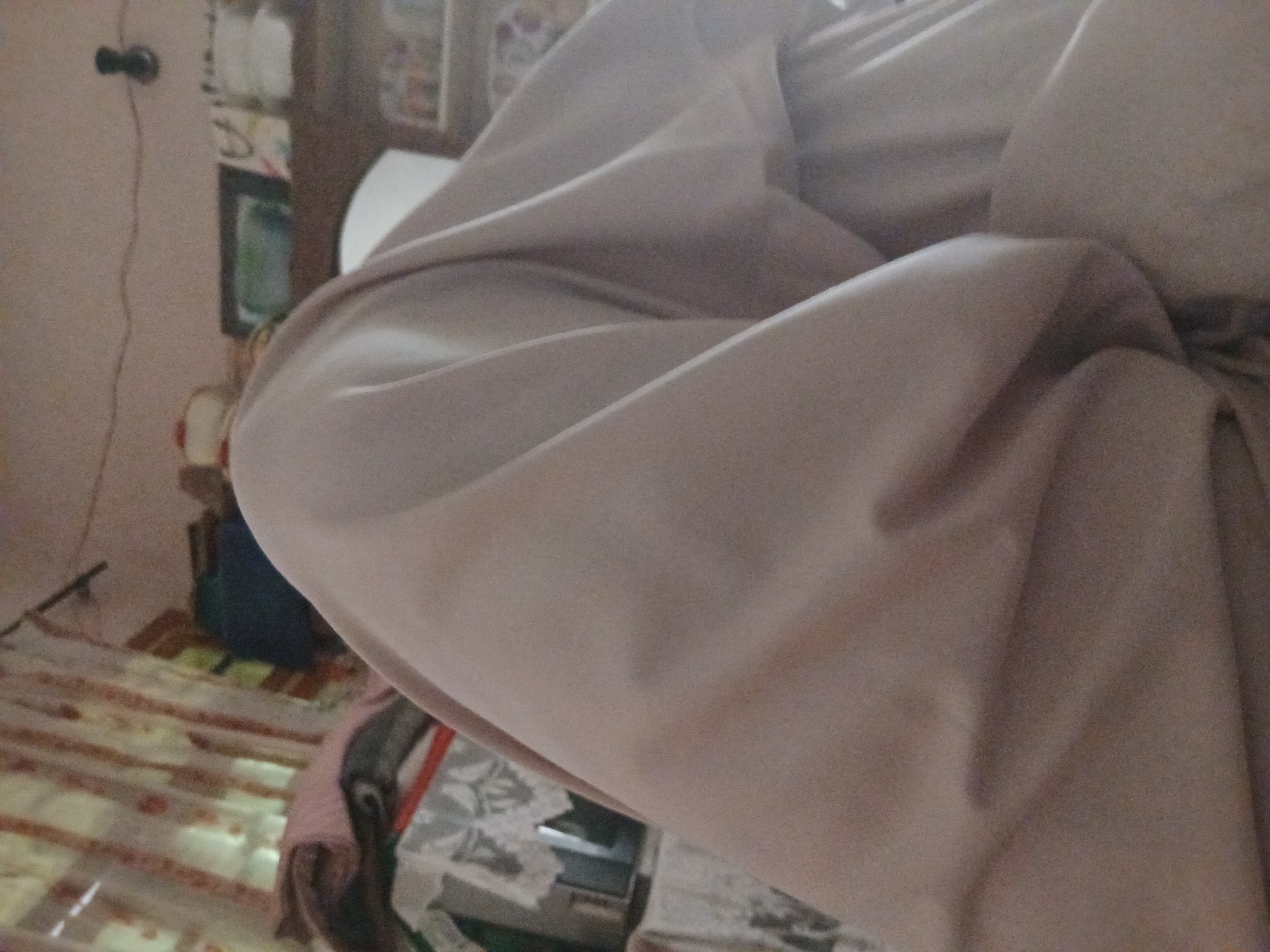 The image depicts an indoor scene with a focus on draped fabric, likely part of a garment or bedding. The fabric is a light, neutral color with soft folds and shadows. In the background, hints of a room are visible, including curtains with a vertical striped pattern, shelves with various objects, and a dark circular object mounted on the wall, possibly a light fixture. The overall impression is of a domestic setting, potentially a bedroom or living area. The image does not provide specific details that would allow for localization to Peshawar City Tehsil, Pakistan.