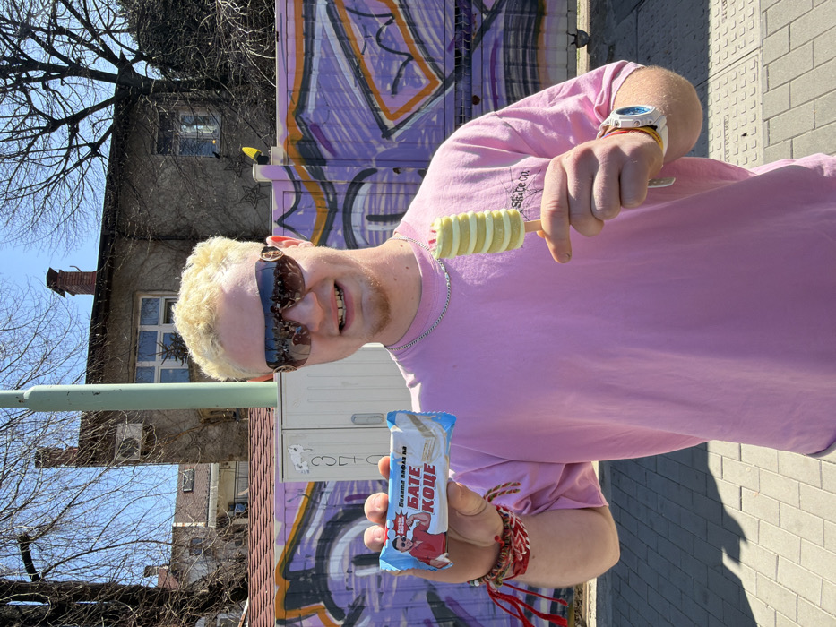 A young man with bleached blonde hair and dark, reflective, wrap-around sunglasses featuring gold-colored emblems stands outdoors on a paved surface in Sofia, Bulgaria, under a clear blue sky. He wears a pink oversized t-shirt, a silver chain necklace, and multiple multi-colored fabric bracelets on his left wrist, with a white digital watch on his right. He faces the camera, smiling, revealing a gap between his front teeth. In his left hand, he holds a light blue packaged rectangular wafer bar, partially obscuring Cyrillic text that includes "ВАЛАТА ВАФЛА на БАТЕ КОЦЕ" and an image of a man. In his right hand, he holds a swirl-shaped, predominantly yellow-white ice cream with a pink-red top accent, on a wooden stick. Behind him, a light purple wall is heavily adorned with abstract graffiti in darker purple and orange-white hues. To the left, a white utility box is visible, marked with what appears to be faint Cyrillic numbers. In the deeper background, several older, two-story buildings with varying facades (stucco, exposed brick, some covered in bare vines) are discernible, alongside bare tree branches and a tall, light green pole. Shadows are cast on the light grey paved ground to the left.