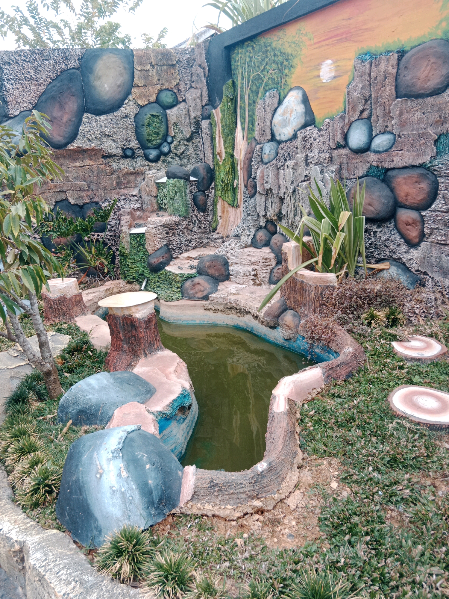 The image shows an artificial pond and rock garden landscape designed to mimic a natural waterfall and mountain scene. The pond is irregularly shaped and filled with murky green water. Surrounding the pond are sculpted rocks of various sizes, some painted to resemble natural stone, and others with mossy green accents. There are also decorative elements resembling tree stumps, some with flat, circular tops, possibly for seating or decorative purposes. Lush green grass and some small plants are growing around the pond and rocks. In the background, a textured wall is visible, painted with a mural depicting a sunset over rocky cliffs and trees. This scene is located in Pualam Sari, Indonesia, specifically in a garden or park setting. There are no people or animals visible in the image.