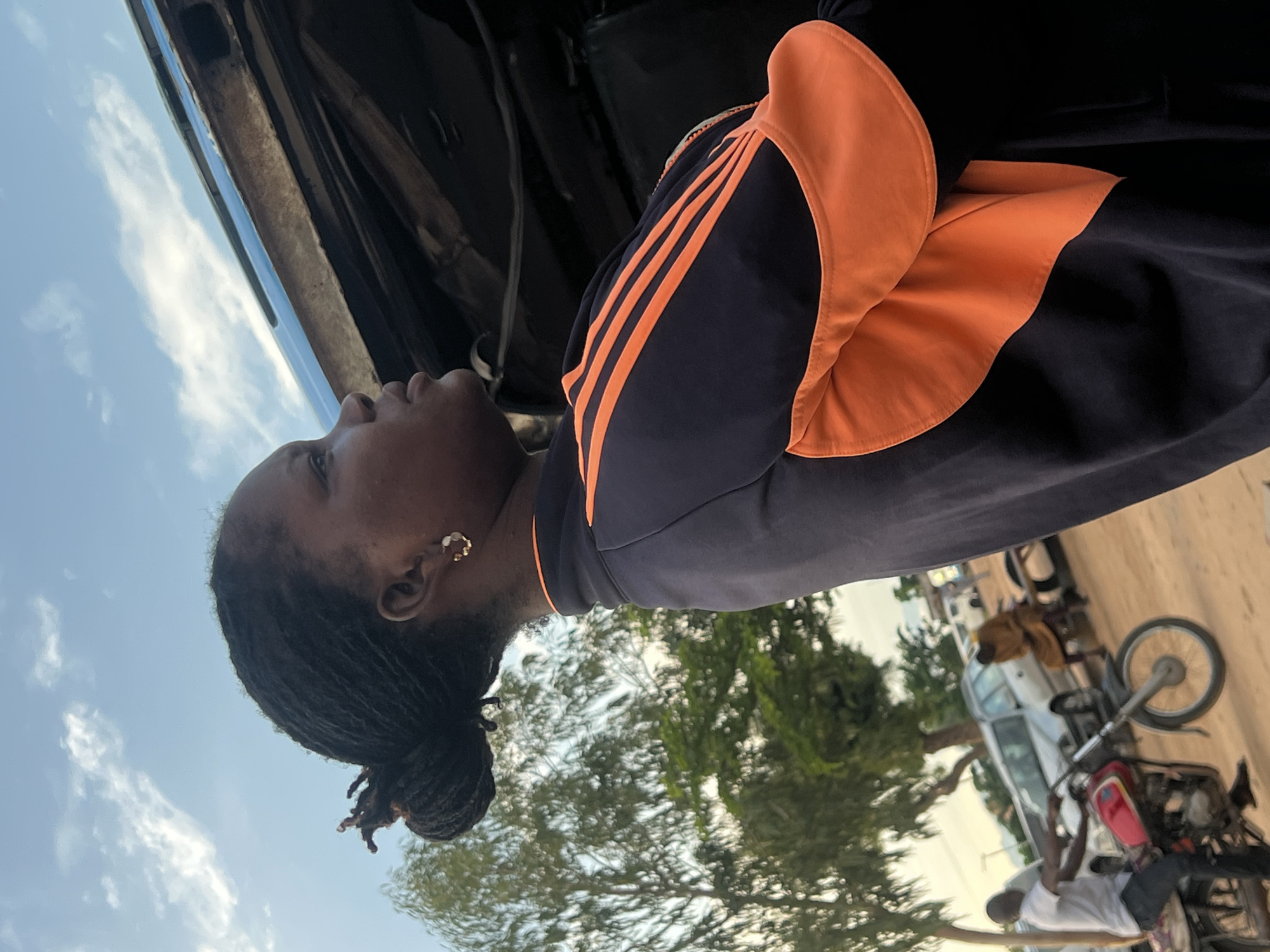 The image depicts an outdoor scene in the city of Gbaza, Nigeria, featuring an adult female in profile on the right side of the frame. She has dark skin, braided hair styled into a bun, and wears a small gold-toned hoop earring with white accents. She is dressed in a dark blue athletic jacket with prominent orange stripes on the sleeve and orange fabric panels on the shoulders/upper arms, indicative of an Adidas brand design. Her gaze is directed upwards and to the right, towards a clear blue sky with scattered white clouds. Behind and above her, a weathered, rusty metal structure with visible blue paint is present, likely a component of an open vehicle. To the left, a man in a light-colored t-shirt and dark trousers is seated on a red motorcycle, facing left, with his feet touching the unpaved dirt ground. Further in the background, a silver sedan is partially visible behind the motorcycle. Another individual, dressed in a light brown garment, is seated on a black tire in the distant background. Green foliage from trees is visible on the left side of the frame and in the midground. The overall environment suggests a semi-urban or rural setting with unpaved roads and visible activity.