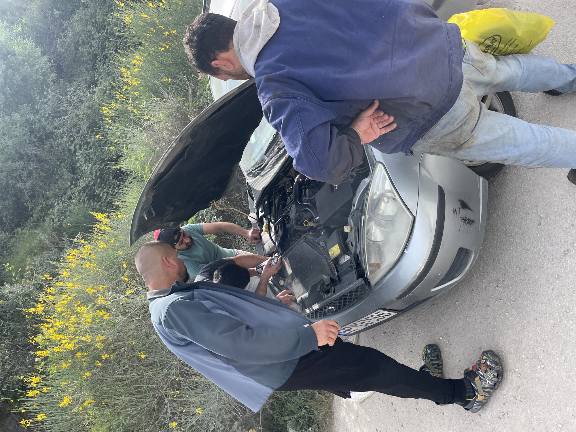 Three men are seen working on a silver car with its hood open. They appear to be focused on the engine. One man is wearing a blue jacket and light blue jeans. Another man is wearing a green t-shirt and a red cap. The third man is wearing a grey and blue jacket and black pants. He is also wearing patterned Crocs. The car is parked on a paved surface next to a bush with yellow flowers. The license plate is visible on the car and reads "BR 00595". The setting seems to be roadside in Čeluga, Montenegro.