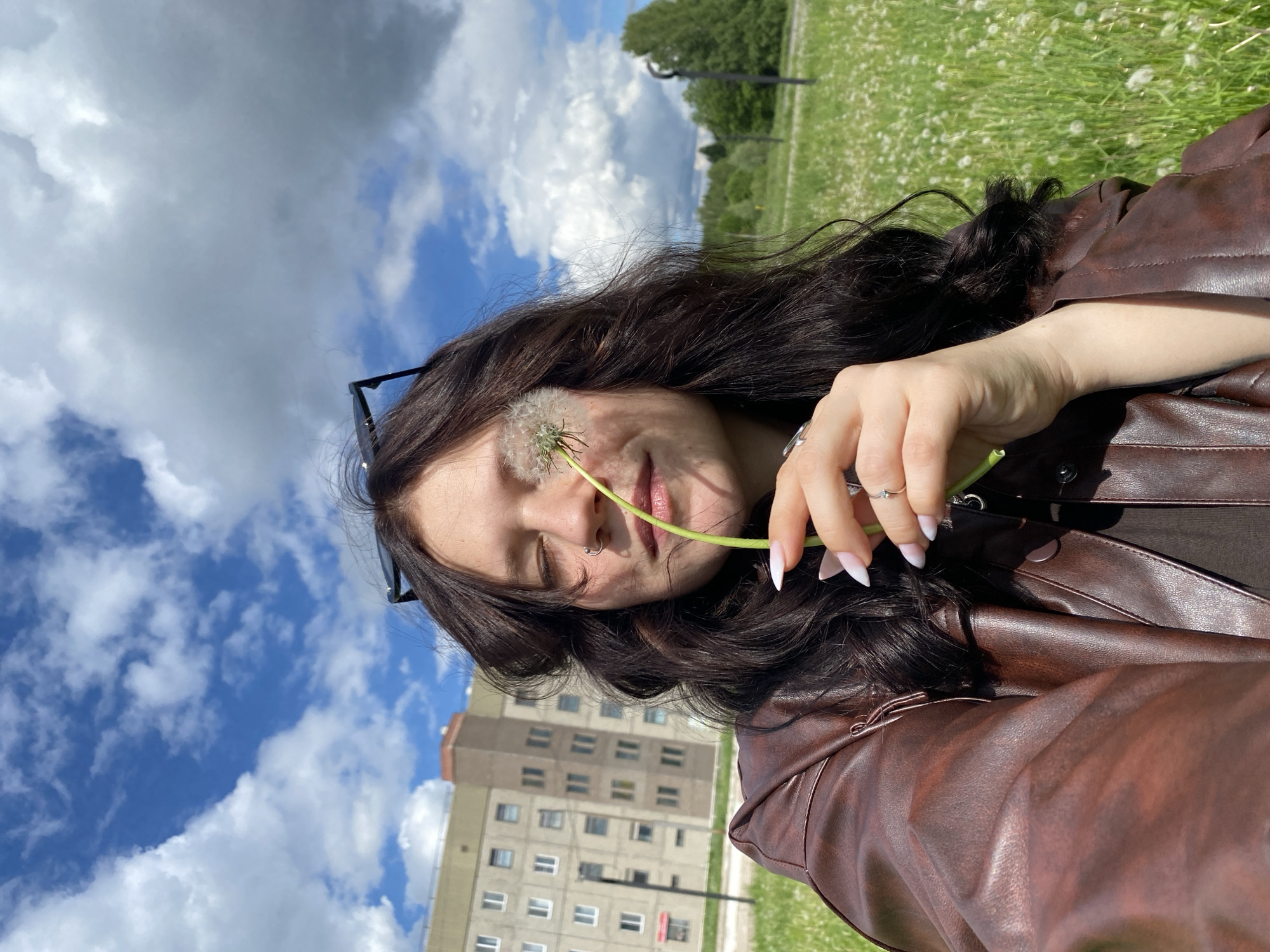 A young woman is pictured in a selfie-style photograph, seemingly taken in a grassy area, possibly a park or field in Марковский, Russia. She is holding a dandelion in front of her face, specifically near her nose. The woman has dark hair and is wearing a brown leather jacket. Sunglasses are perched on her head. Her nails are manicured with a white tip design. Behind her is a cloudy blue sky and a multi-story building. The ground is covered in green grass and small white flowers, possibly daisies or dandelions. In the distance there are trees and an art installation.