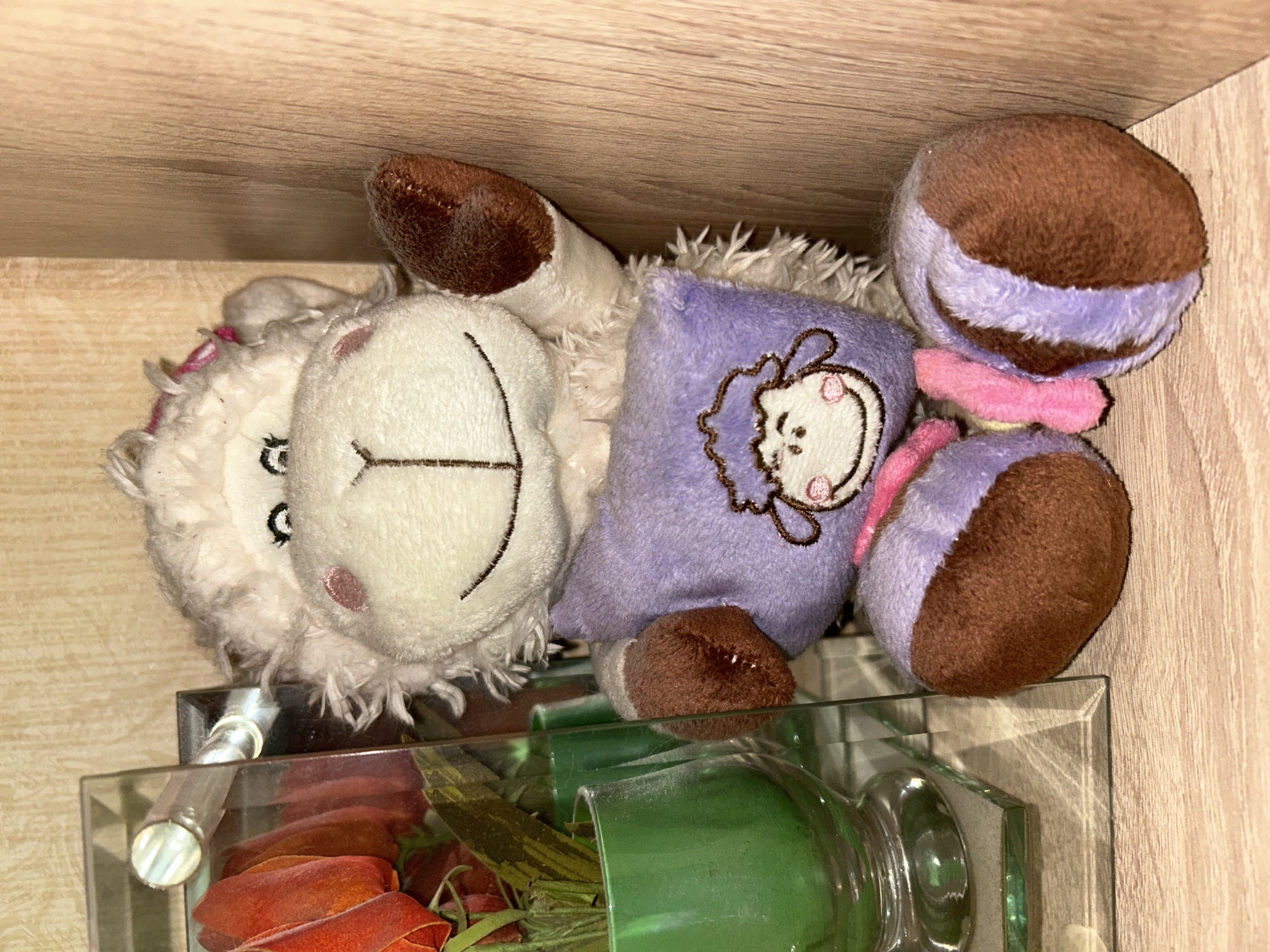 A fluffy, white stuffed sheep toy is seated on a wooden shelf. The sheep has a large, round face with embroidered eyes and a smiling mouth. It is wearing a purple vest with a smaller embroidered sheep face on it, and pink accents. Its arms and legs are brown and fluffy. To the left of the sheep is a clear glass display case containing artificial red flowers in a green vase. The background consists of a textured beige wall and a light brown wooden shelf. The scene appears to be indoors, likely in a residential setting.