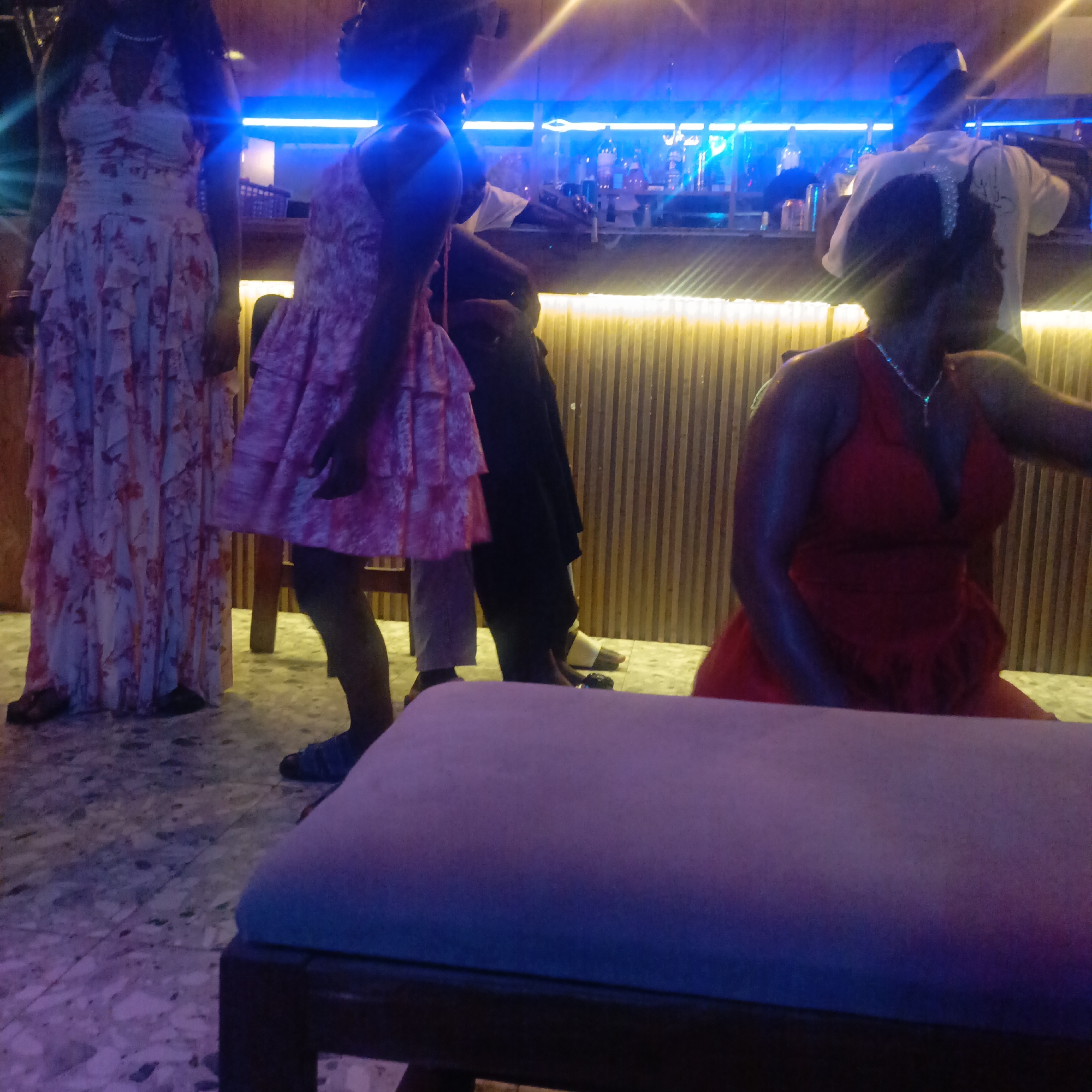 The image shows a dimly lit indoor scene, likely a bar or lounge, in Akwanga, Nigeria. Three women are visible in the foreground. The first woman stands on the left and wears a long, floral dress. The second woman, standing closer to the center, is dressed in a short, pink floral dress. A third woman is seated, wearing a red dress. Additional patrons and a bartender are visible in the background near the bar, which is lined with bottles. A padded ottoman-style seat is in the extreme foreground of the image. The lighting features blue hues. The flooring appears to be tile.