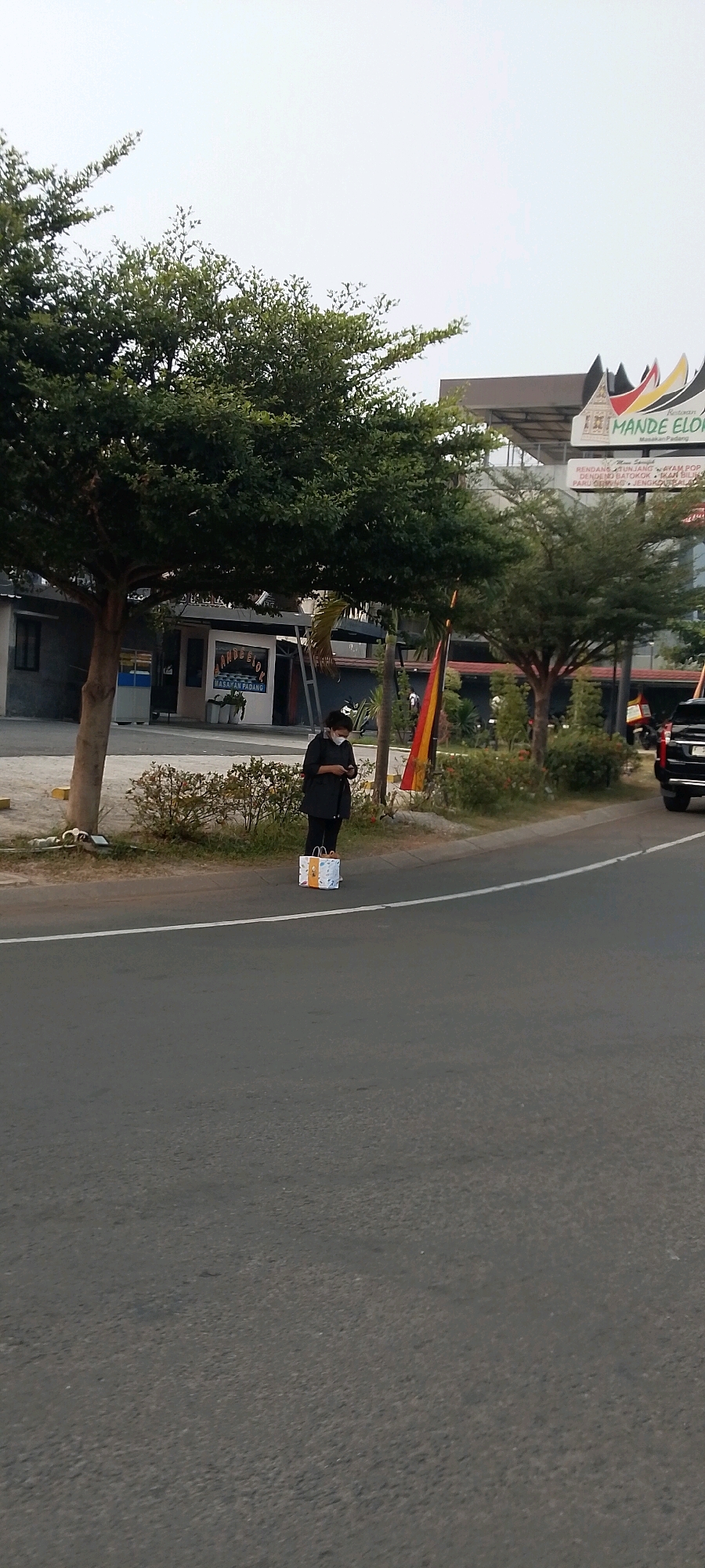A person wearing a black jacket, black pants, and a white face mask stands on the side of a road in Gunung Putri, Indonesia. The individual is looking at their phone. In front of them, on the ground, is a white shopping bag with orange and yellow accents. Behind the person, there are trees, a building with a sign that reads "Mande Elok Masakan Padang", and a black car parked on the side of the road. The road has a white line marking. The sky is overcast.
