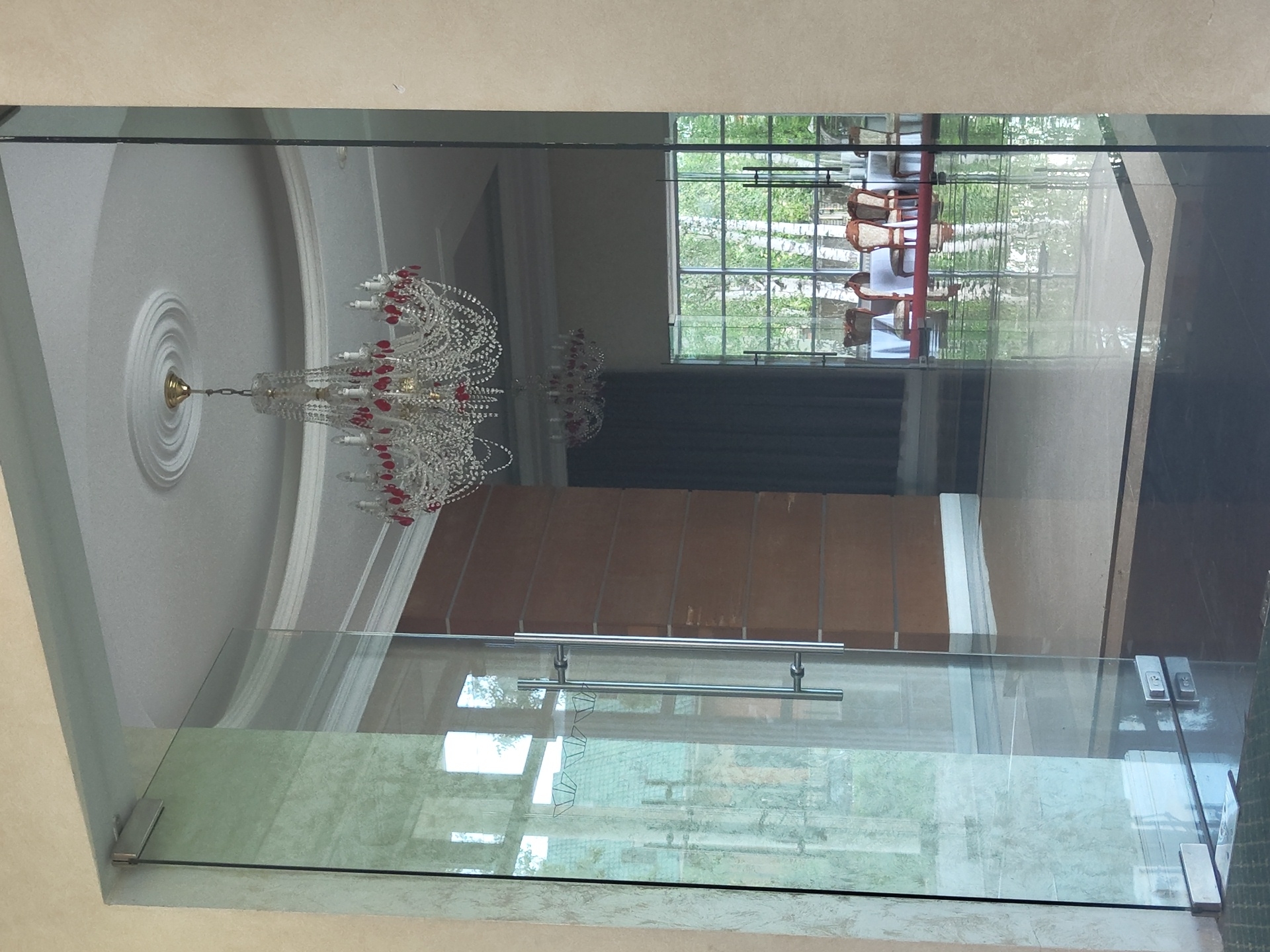 The media file shows a reflection in a glass enclosure. In the reflection, a chandelier with crystal prisms and red accents hangs from a white ceiling with ornate molding. Beyond the chandelier, a window reveals a lush green outdoor scene with trees. Inside the reflected space, there is a table and chairs, suggesting a dining or seating area. The interior also features dark fabric drapes and what appears to be a wall with vertical wooden panels. The foreground of the media file is dominated by the glass enclosure itself, with a metal railing and supporting fixtures. The glass has a slightly textured or frosted lower section. The overall impression is of an indoor space with a view of nature. The lighting suggests it could be daytime, with natural light filtering through the window. No people or specific activities are directly visible, only the static elements of the reflected environment and the glass structure.