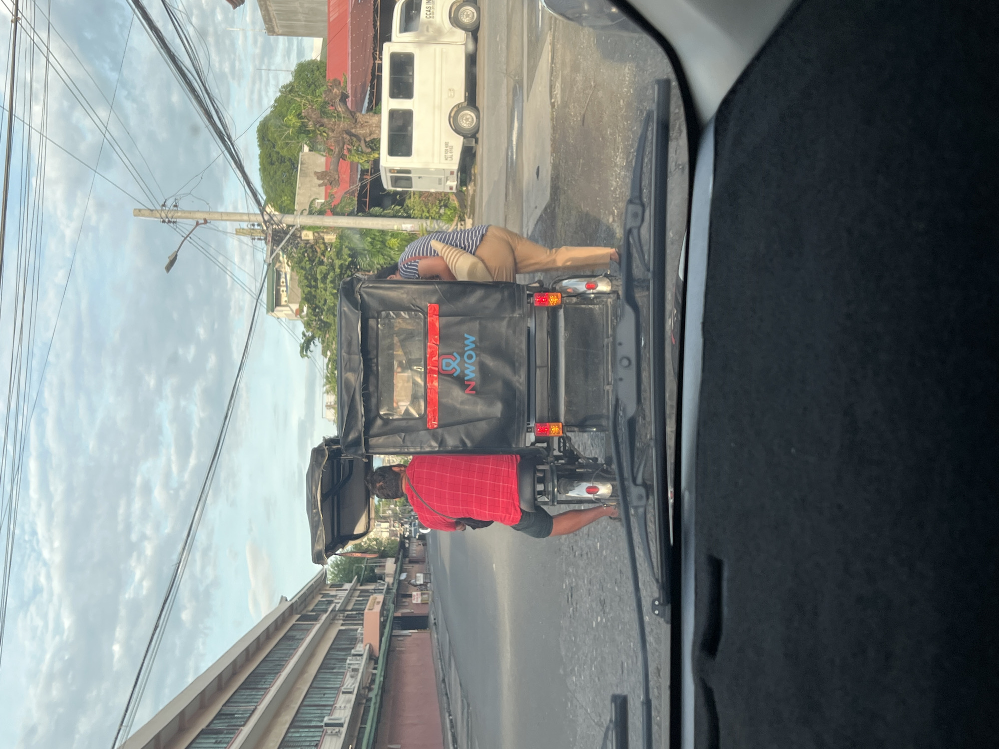 The image shows a street scene in Davao City, Philippines. A tricycle is in the foreground, partially obscured by a vehicle interior (possibly a car). The tricycle has the logo of a company called "NWOW" on the back. One man in a red shirt is riding the tricycle, and another man, wearing a striped shirt and khaki pants, is looking at the engine. Behind the tricycle, there is a white van and a multi-story building. Power lines are visible overhead. The sky is cloudy.