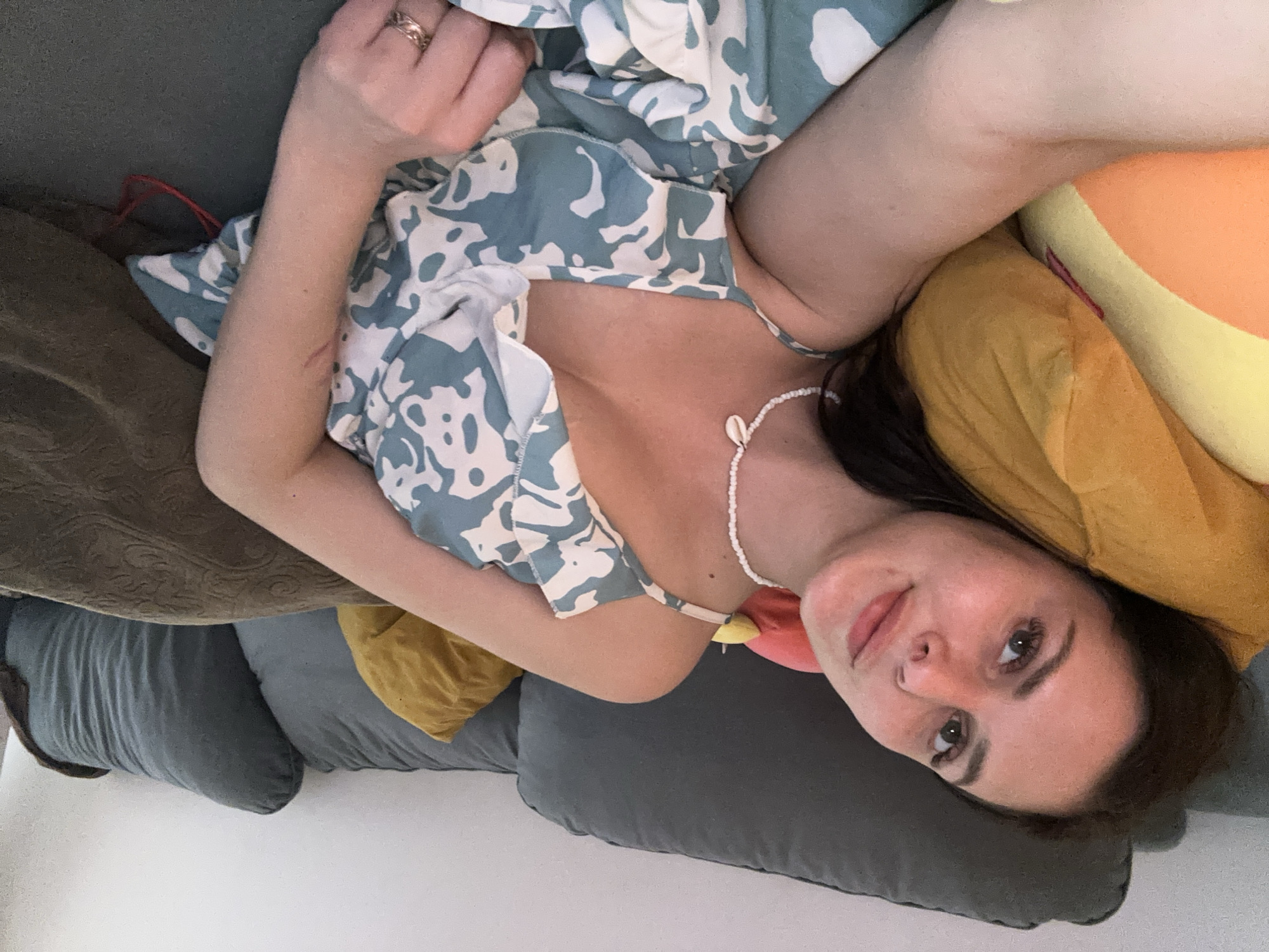 A young woman with brown hair and fair skin is lying on a gray couch, propped up by several pillows. She is wearing a light blue and white patterned dress and a shell necklace. The woman is looking towards the camera with a slight smile. In the background, there are several pillows on the couch, some of which are yellow and orange. A dark blanket is also visible on the couch. The setting appears to be an indoor living room.