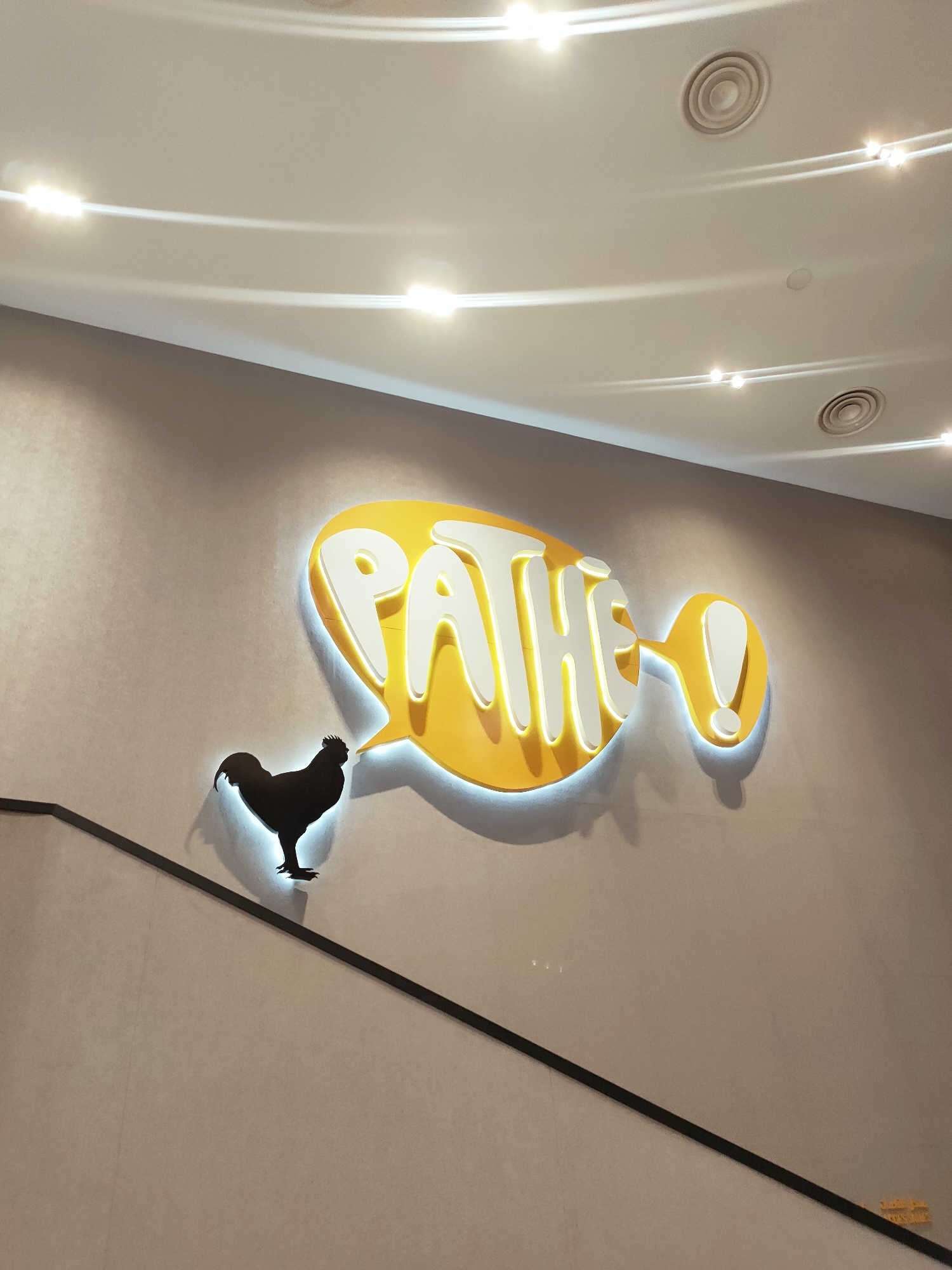 In the city of Cebalat, Tunisia, a dimly lit hallway features a grey textured wall with a black diagonal railing. Mounted on the wall is a large, illuminated yellow speech bubble sign. The sign displays the word "PATHE!" in white, backlit lettering. To the left of the speech bubble, a black silhouette of a rooster stands on a black surface, casting a subtle shadow illuminated by the sign's glow. The ceiling above is visible, with recessed circular vents and curved, illuminated light fixtures.