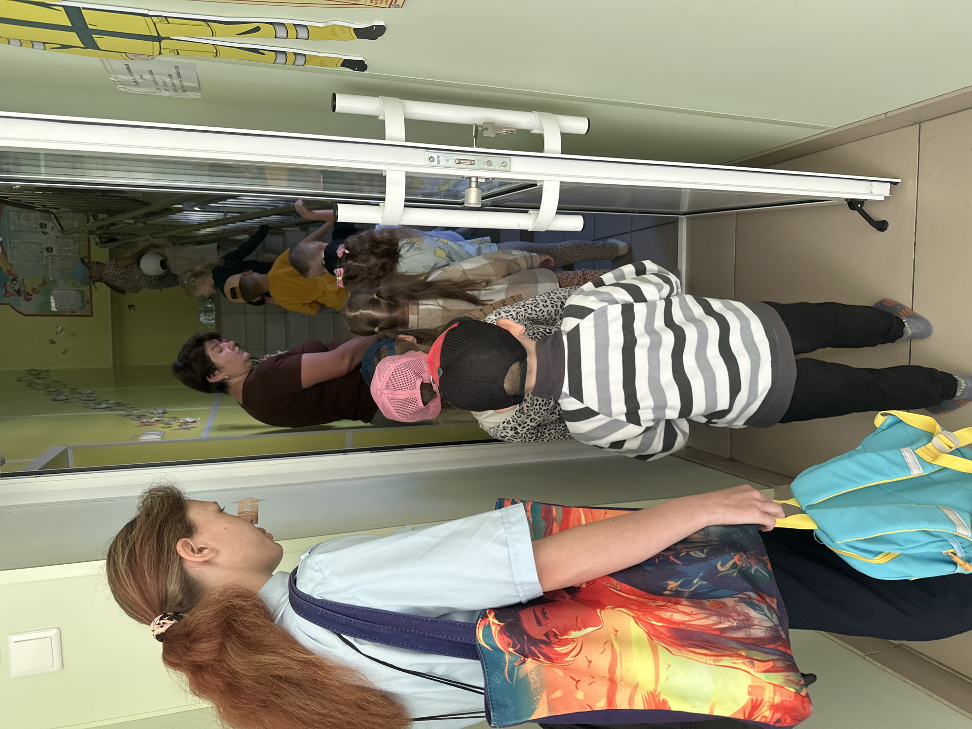 A group of children and an adult are standing in a hallway, partially obscured by a glass door. On the left, a woman with reddish-brown hair pulled into a ponytail is wearing a light blue shirt and carrying a large tote bag with a graphic print. Beside her, to the right, a child wearing a black and grey striped sweater and black pants is walking away from the camera. Behind this child, more children are visible, some wearing hats. Further into the hallway, a woman wearing a dark brown shirt is looking down, and a set of stairs is visible. The hallway walls are painted a light green. A yellow graphic of a figure in a uniform is attached to the wall on the far right. The location is identified as Novoaltaysk, Russia.