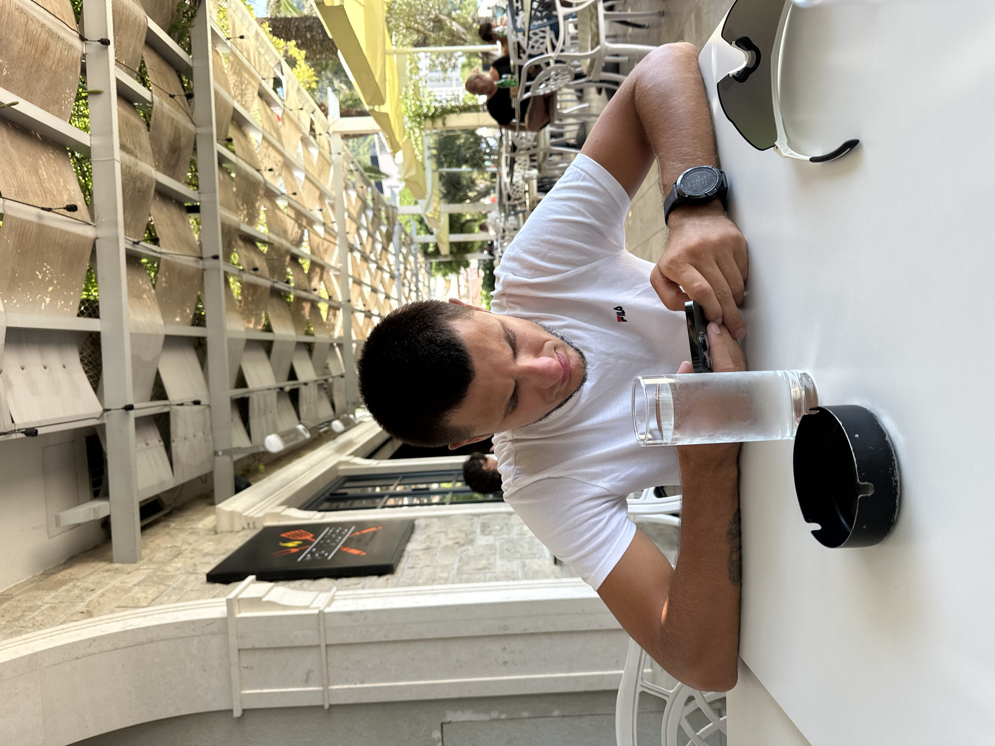 The image shows a young man sitting at a white table outdoors, in Bečići, Montenegro. He is wearing a white t-shirt with a red FILA logo and is looking down at his smartphone. On the table in front of him are a glass of water, a black ashtray, and a pair of white sunglasses. In the background, there are other tables and chairs, some with people seated, and a canopy overhead supported by white beams. The architecture of the building behind him features light-colored stone and a large archway. The environment appears to be a restaurant or cafe with an outdoor seating area, suggested by the presence of umbrellas and trees visible further in the background, indicating a sunny day.