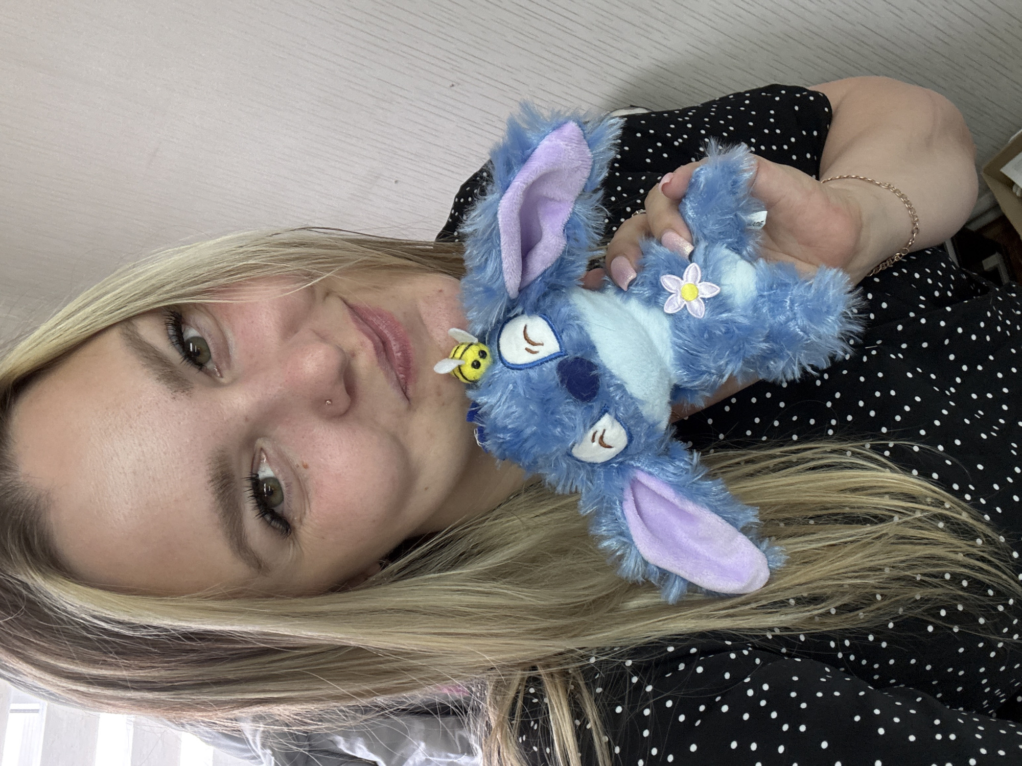A young woman with blonde hair is holding a blue plush toy with long ears. The toy has a bee and a flower attached to it. The woman is wearing a black and white polka dot shirt and a gold bracelet. Her hair is partially covering a gray fabric in the background, and she has a nose piercing. The location is not identifiable as Noviy, Russia, based on the provided image.