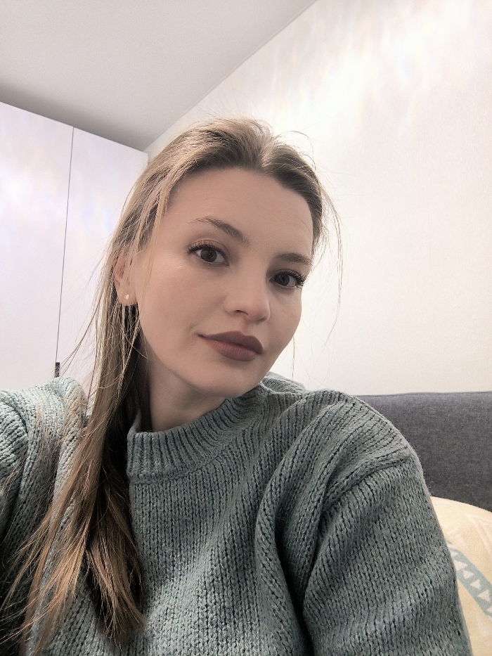 A woman with light brown hair and fair skin faces the camera. She wears a sage green knit sweater. Her makeup consists of dark lipstick and defined eyebrows. She has small stud earrings. Behind her is a white wall and part of a gray sofa. The lighting is soft and diffused, highlighting her features.