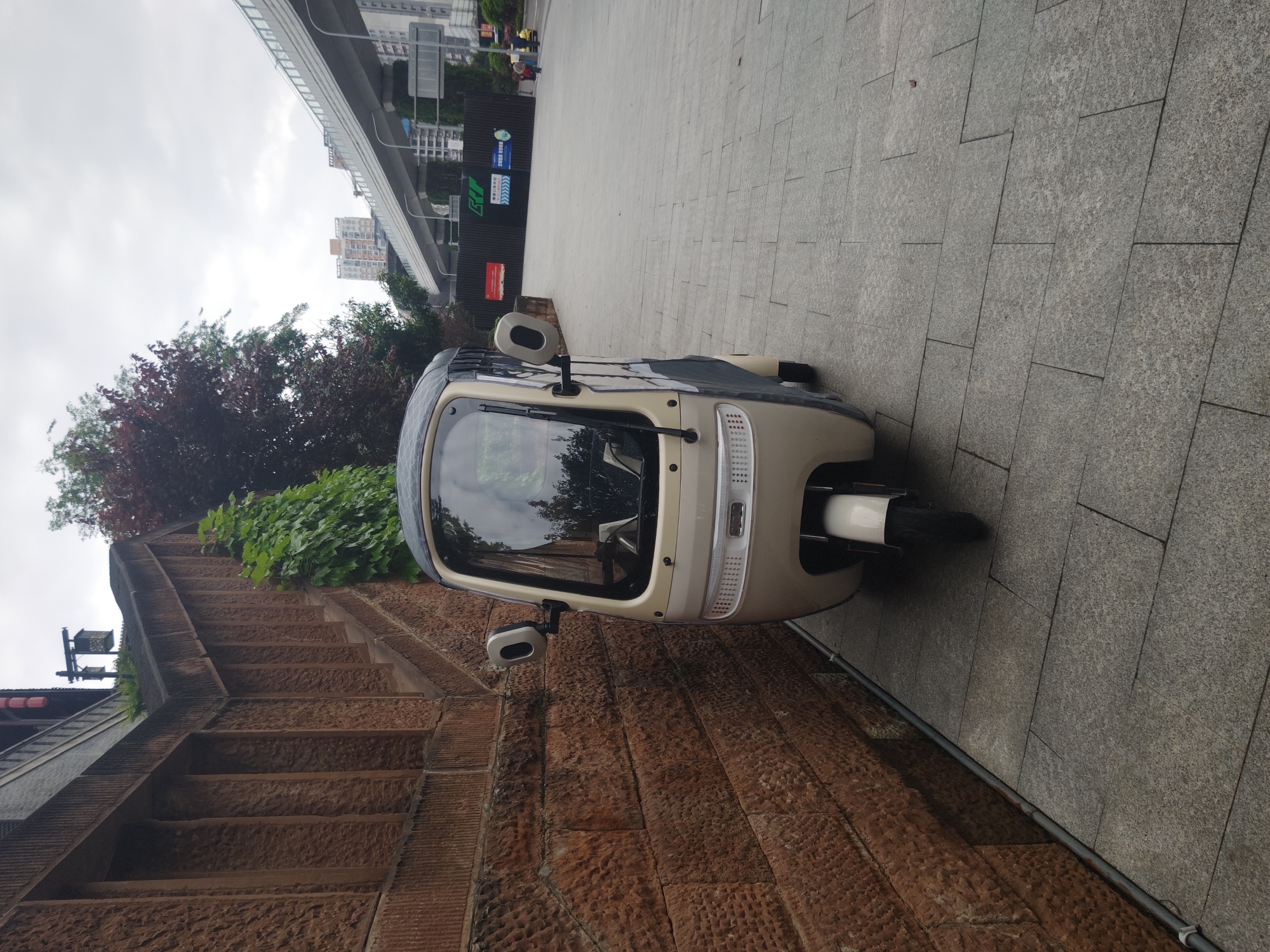 A motorized tricycle is parked on a stone-tiled walkway adjacent to a brown stone wall with stairs. The tricycle is light beige and black with a windshield and two side mirrors. Above the wall is greenery, including trees and vines. Beyond the tricycle, the walkway extends into the background, where there is a building, signs, and a pedestrian overpass. The sky is overcast. The location is in the Shapingba District, China.