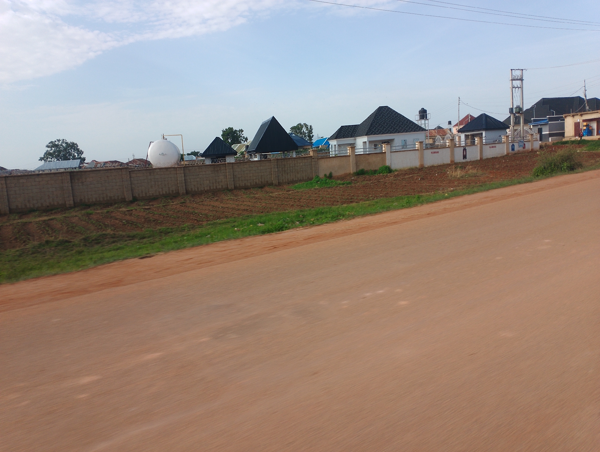 The image depicts a street view in Kwang, Nigeria, with a row of houses and a large wall in the background. The houses have modern architecture with dark, pitched roofs. Some of them are painted white, while others have light-colored exteriors. A large, spherical white tank is visible behind the wall, possibly related to water storage or some industrial process. A utility pole with electrical equipment stands near the houses. The foreground is dominated by a reddish-brown dirt road, suggesting an unpaved or newly constructed area. Green grass and some dry soil are visible beyond the wall, bordering the houses. The sky is partly cloudy with a bright, possibly daytime sky. No people are clearly visible in the image. The overall impression is one of development and construction in a semi-rural or suburban setting. There is no visible text in the image.
